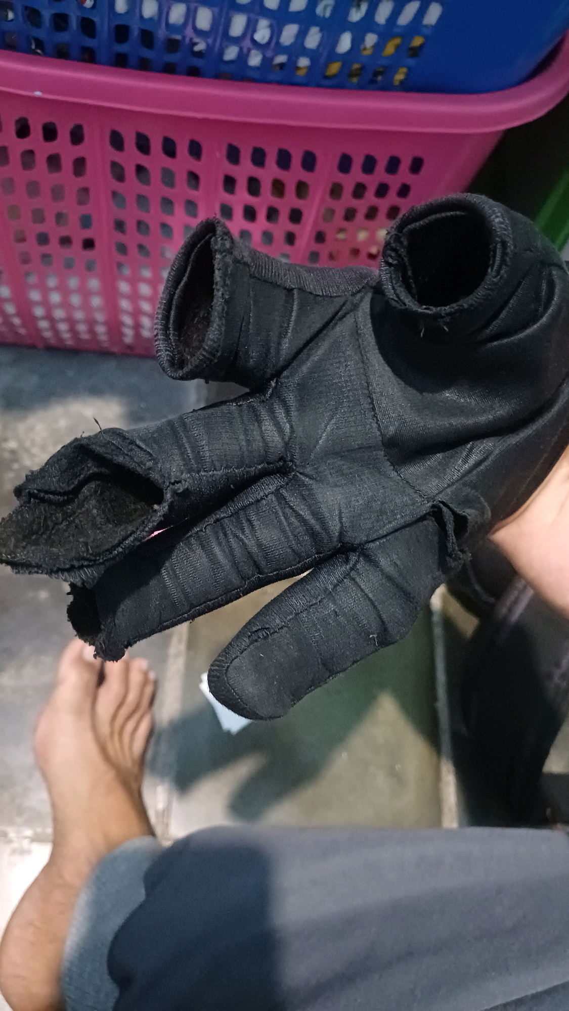 A pair of torn black gloves is held in a person's right hand, which is positioned in the foreground. The gloves appear to be made of a synthetic material and show significant wear and tear, with rips and exposed stitching on the fingers and thumb. In the background, two plastic laundry baskets, one pink and one blue, are stacked. The floor appears to be concrete. A bare foot and lower leg of the person holding the gloves are visible in the bottom left of the frame. The scene is indoors, likely in a residential setting, and the lighting suggests it is daytime. The specific location of Kowangan, Indonesia, cannot be determined from the visual information provided.