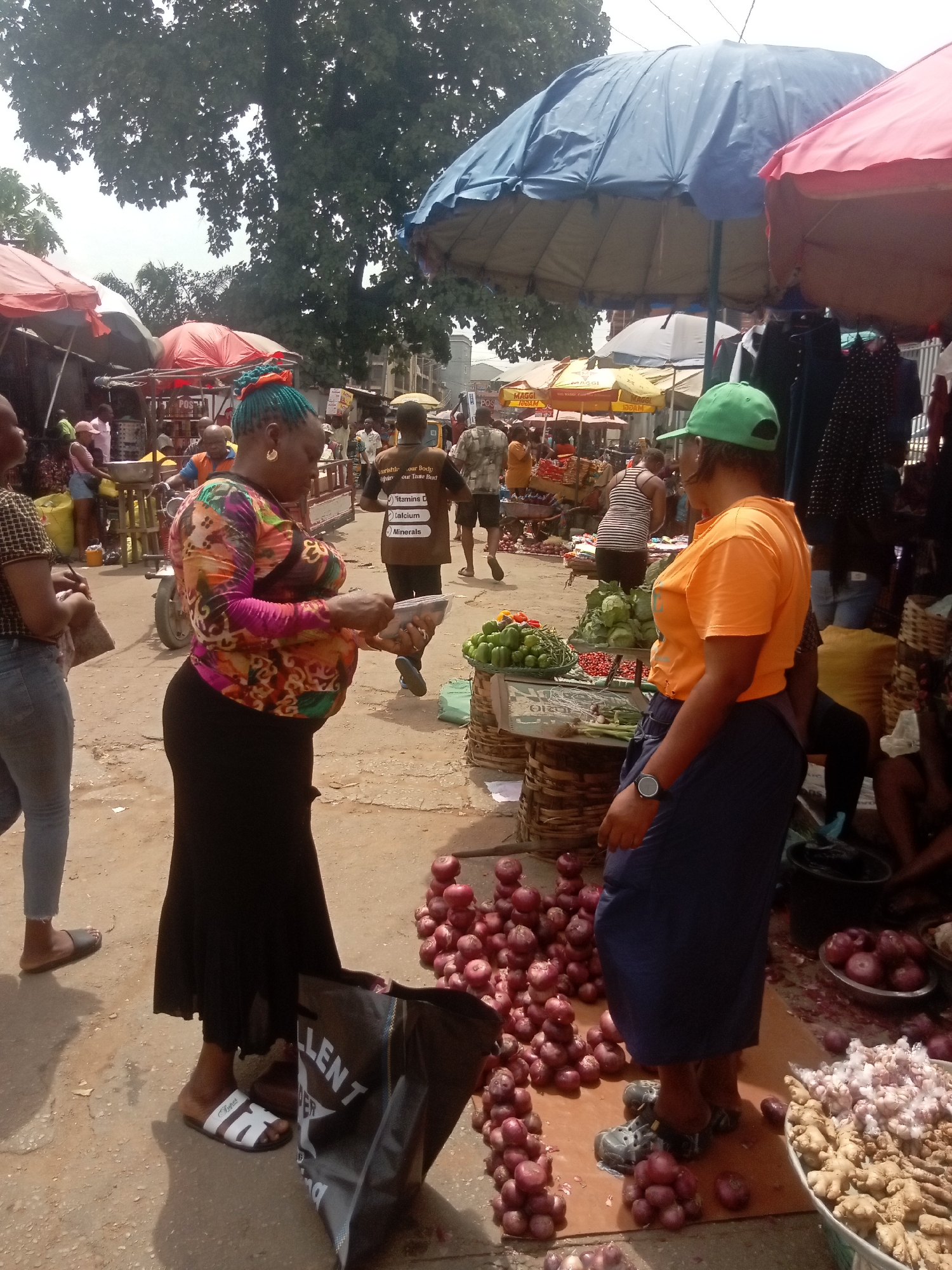 In a bustling open-air market in Onitsha, Nigeria, a woman with dyed teal hair and a colorful blouse is seen paying a vendor. The vendor, wearing an orange shirt and green baseball cap, stands beside her produce: an assortment of bell peppers, cabbage, and tomatoes. A pile of red onions is scattered on the ground between them, along with ginger and garlic. Other market vendors, stalls covered by colorful umbrellas, and customers are visible in the background. A black shopping bag rests at the feet of the woman making the purchase.