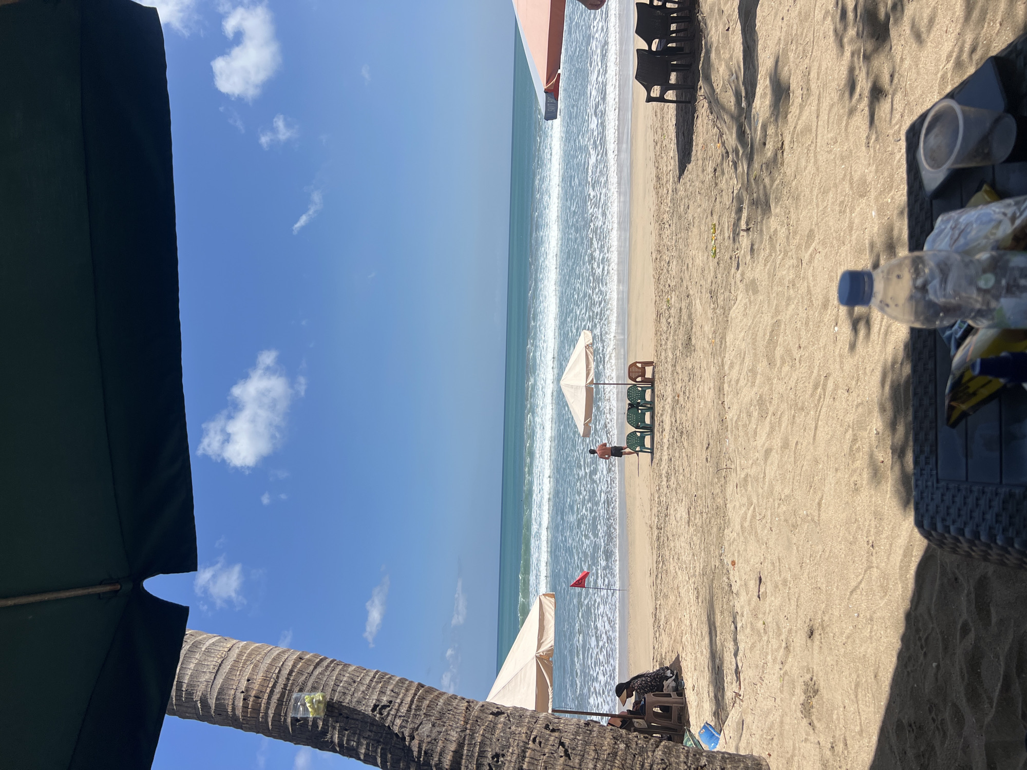 The image depicts a beach scene in Kuta, Indonesia, with the ocean and a clear blue sky in the background. In the foreground, there is a table with a plastic water bottle, a clear plastic cup, and a smartphone on it. Partially visible shadows suggest there are umbrellas providing shade.
Further into the scene, the sandy beach is dotted with beach chairs and umbrellas. A red flag is planted in the sand, possibly indicating swimming conditions. A man in swim trunks stands near a set of green chairs under an umbrella, facing the ocean. To the left, under another umbrella, a person wearing a hat is seated, appearing to be at a table. A palm tree trunk is visible on the far left, with a small clear plastic bag containing items attached to it. Waves are breaking on the shore, and there are a few distant figures in the water. The overall impression is a typical beach day scene with amenities for visitors.