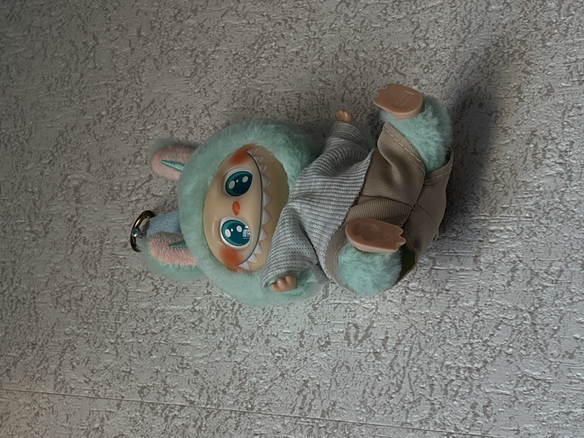 A plush toy with a light blue, fuzzy hood and pink bunny ears is attached to a grey, textured wall. The toy has a light beige face with large, blue, cartoonish eyes and rosy cheeks. It has a wide smile with visible white, jagged teeth. The toy wears a light grey, ribbed sweater and beige pants. Its arms and legs are light blue and fuzzy. The toy is positioned as if sitting or lying down, with its legs extended to the right. A metal keychain ring is attached to the top of its hood. The background is a plain, textured wall, typical of interior walls. The image was taken in Genemuiden, Netherlands.