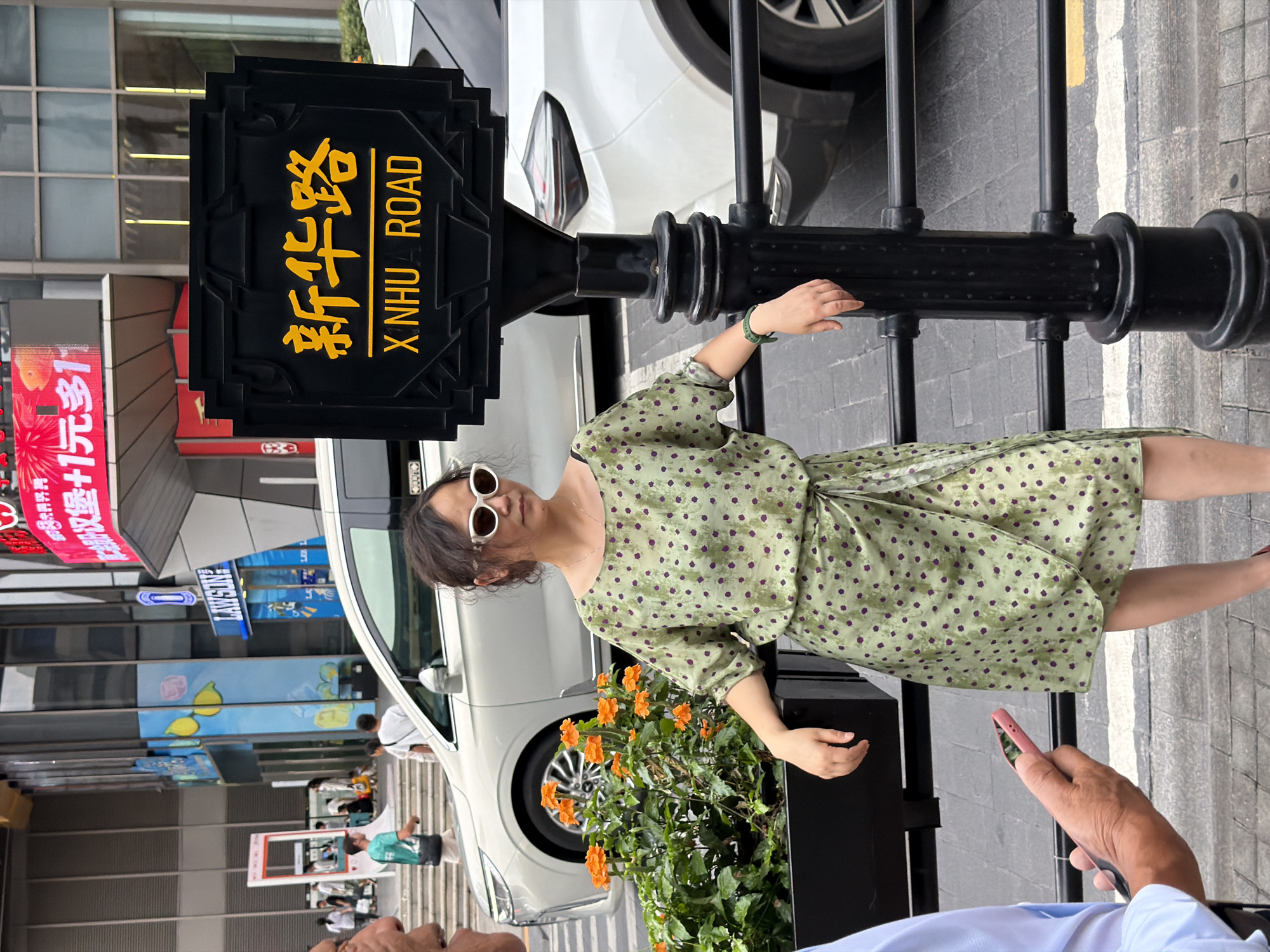 The image shows a street scene in Yuzhong District, China, featuring a woman standing next to a street sign that reads "XINHUA ROAD" in English and Chinese characters. The woman is wearing a light green dress with a polka dot pattern and white sunglasses. She is leaning on a black metal railing. In the background, there are buildings, cars, and other pedestrians. The street sign is black with yellow text and is mounted on a black post. There are orange flowers in a planter to the left of the woman. A man's hand holding a pink smartphone is visible in the lower left corner of the image.