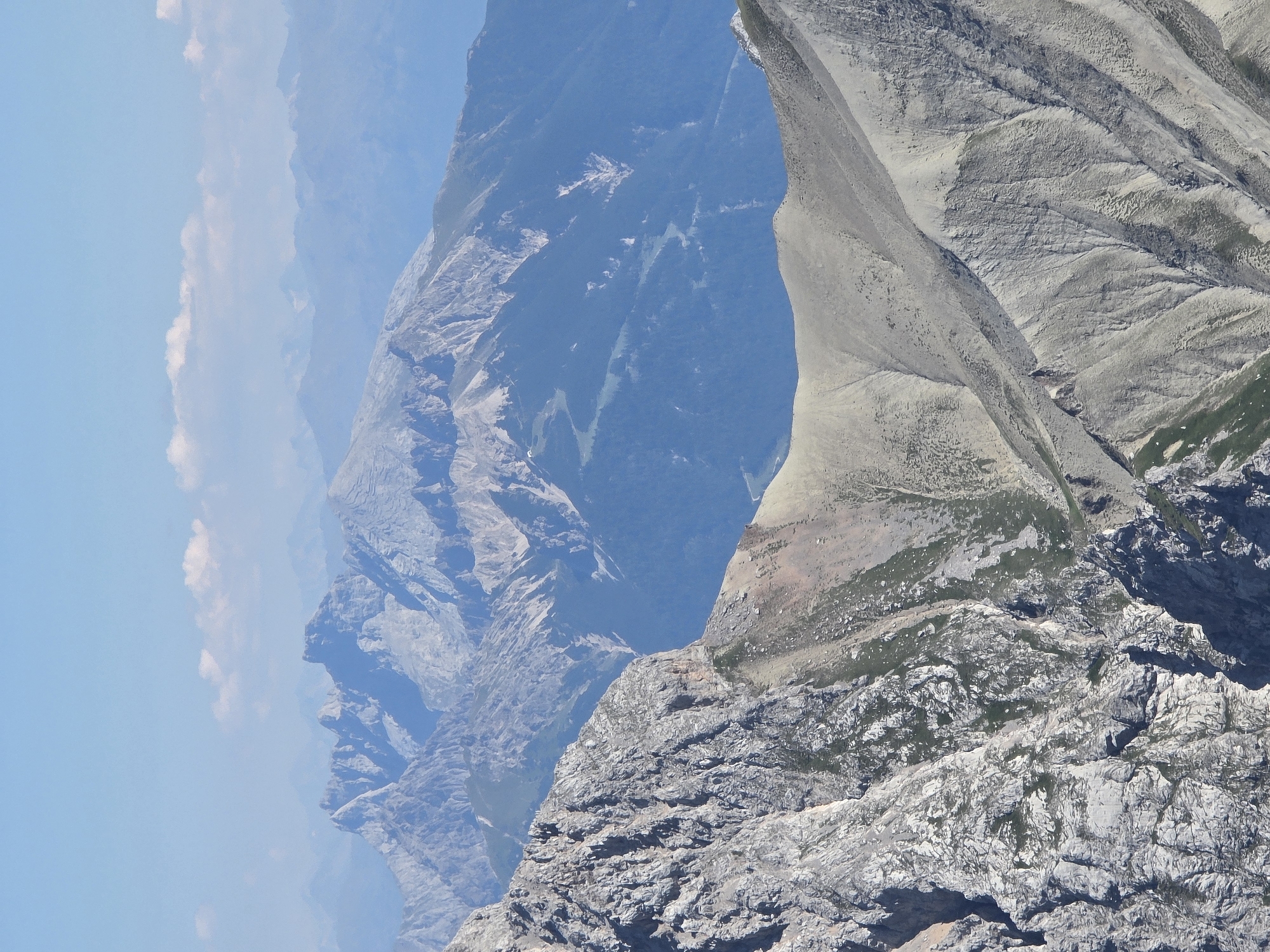 This is a vertical, medium-shot, eye-level, outdoor photograph of a vast mountain range under a clear blue sky with a few scattered clouds. The foreground features a steep, rocky, grey mountain slope with sparse green vegetation. In the middle ground and background, successive layers of mountain ranges recede into the distance, becoming progressively bluer and hazier due to atmospheric perspective. The mountains exhibit sharp, jagged peaks and undulating ridges. The scene is devoid of any people or man-made structures, suggesting a remote, natural environment. The location is identified as the mountains near Garmisch-Partenkirchen, Germany, a popular region for alpine scenery and outdoor activities.