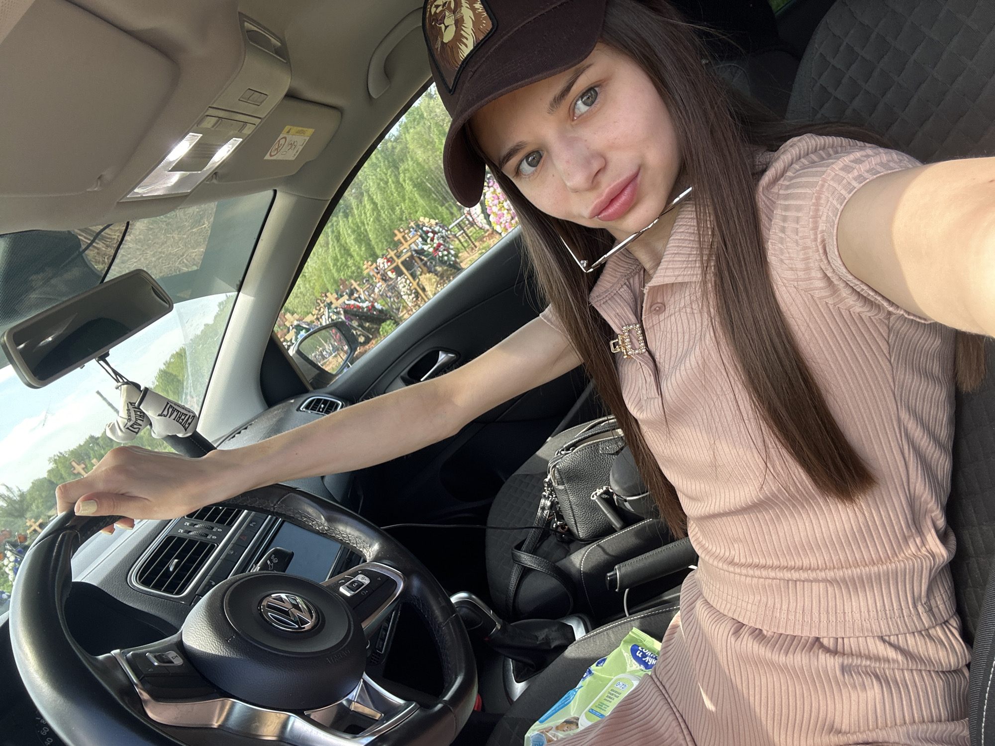 A woman is in the driver's seat of a car, holding the steering wheel. She is wearing a brown baseball cap with a lion design and a peach-colored collared shirt. A black purse is on the passenger seat, and baby wipes are visible as well. Outside the car window, a landscape of green trees and many crosses indicates a graveyard, identified by the location metadata as likely being in Izhevsk, Russia.