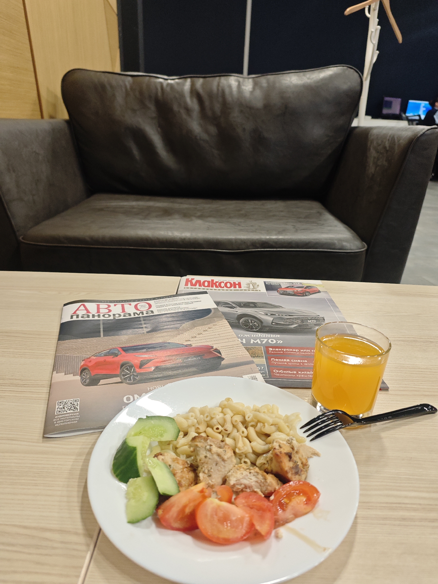 The scene is an indoor setting, possibly a lounge or waiting area, evident from the presence of a brown leather-like armchair in the background. A low table is in the foreground, on which a white plate holds a meal composed of macaroni, grilled chicken or other meat, sliced tomatoes, and cucumber. Next to the plate is a glass of orange juice and a black fork. Beneath the plate and glass, there are two Russian magazines, identified by their titles "ABTO панорама" and "Клаксон," both related to cars, suggesting the location could be a car dealership or service center. The OCR output from the image suggests some text related to car features and brands, which reinforces the car theme. In the background, the presence of a coat rack and a partial view of a person in front of computer screens imply a functional space. Without further information, it's impossible to definitively confirm the location in Bolshoye Savino, Russia.