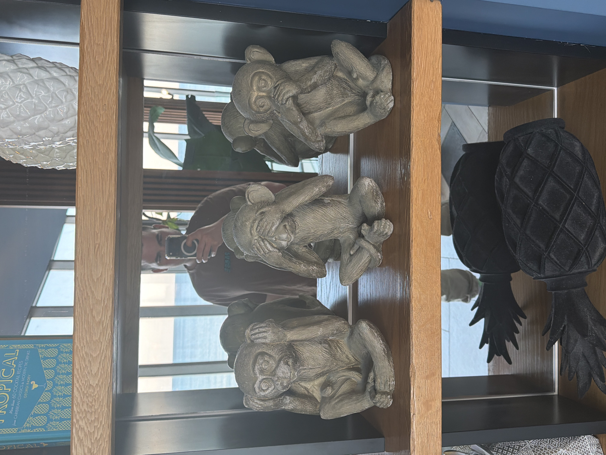 The image, rotated 90 degrees counter-clockwise, captures a decorative shelving unit. When viewed in its correct upright orientation, the unit features light wooden vertical supports and dark horizontal shelves with mirrored backings.
On the uppermost visible shelf, a white textured pineapple-shaped decorative object is placed on the left side. On the middle shelf, three gray monkey figurines are displayed, depicting the classic 'see no evil, hear no evil, speak no evil' poses. Two monkeys are on the left section of this shelf, and one is on the right section, separated by a wooden support. On the bottom shelf, a blue book is positioned on the left side, while two dark, textured pineapple-shaped objects are arranged on the right side.
The mirrored back of the shelves prominently reflects the person taking the photograph, visible holding a smartphone. The reflection also reveals a large window in the background, offering a view of an urban landscape with buildings and a bright blue sky, suggesting it is daytime. This setting is consistent with an indoor location in Varna, Bulgaria.
The visible text on the blue book reads: "TROPICAL", "More than 60 COCKTAIL RECIPES", "CARIBBEAN CLASSICS & MODERN TIKI DRINKS", and "GEORGI RADEV".