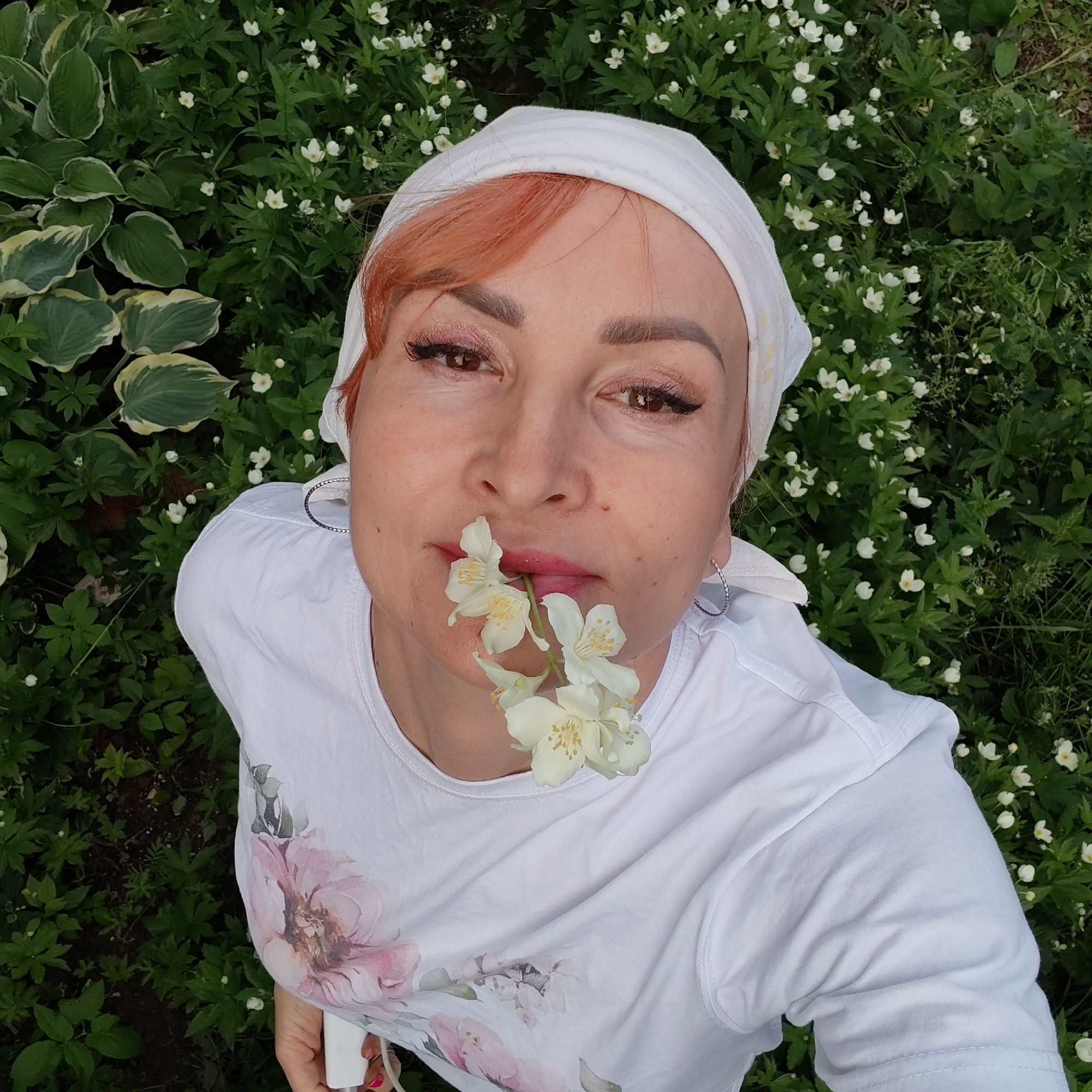 A woman with auburn hair, wearing a white headscarf and a white t-shirt with a floral print, holds white jasmine flowers in her mouth. She is outdoors, with lush green foliage and small white flowers surrounding her. The setting appears to be a garden or park, possibly in the city of Chaikovsky, Russia. The woman is looking directly at the camera.