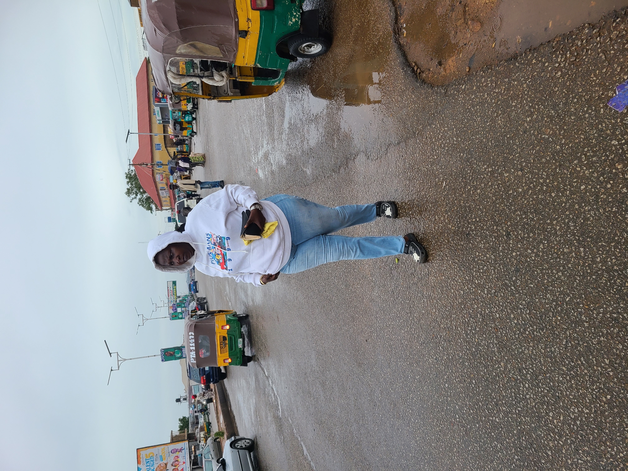 A person stands on a wet, asphalt road in Gura Top, Nigeria. The person wears a white hoodie and blue jeans, holding a phone and cloth. The road is wet and reflective, indicating recent rain. Multiple vehicles, including green and yellow tricycles, are parked or driving on the left side of the road. Buildings and streetlights are visible in the background. The sky is overcast.