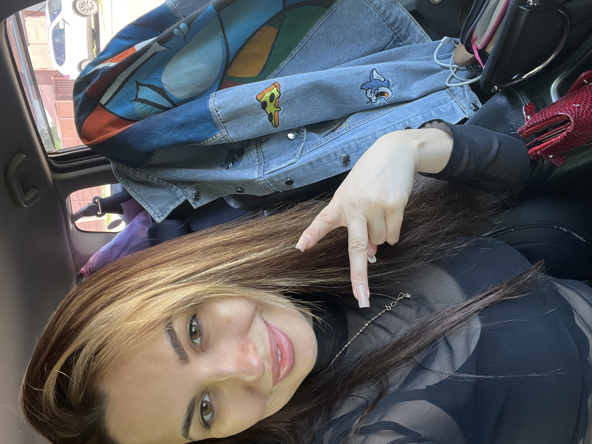 The image shows a woman seated inside a vehicle. The location is specified only as Unknown, Unknown. She is smiling and pointing downward with her right hand. She is wearing a black top with a mesh overlay and a delicate chain necklace. Her hair is long, brown with blonde streaks. Behind her are various items including a blue denim jacket with patches and designs, a black bag, and a red object. The vehicle has a sunroof, through which a glimpse of a white car and a building can be seen.