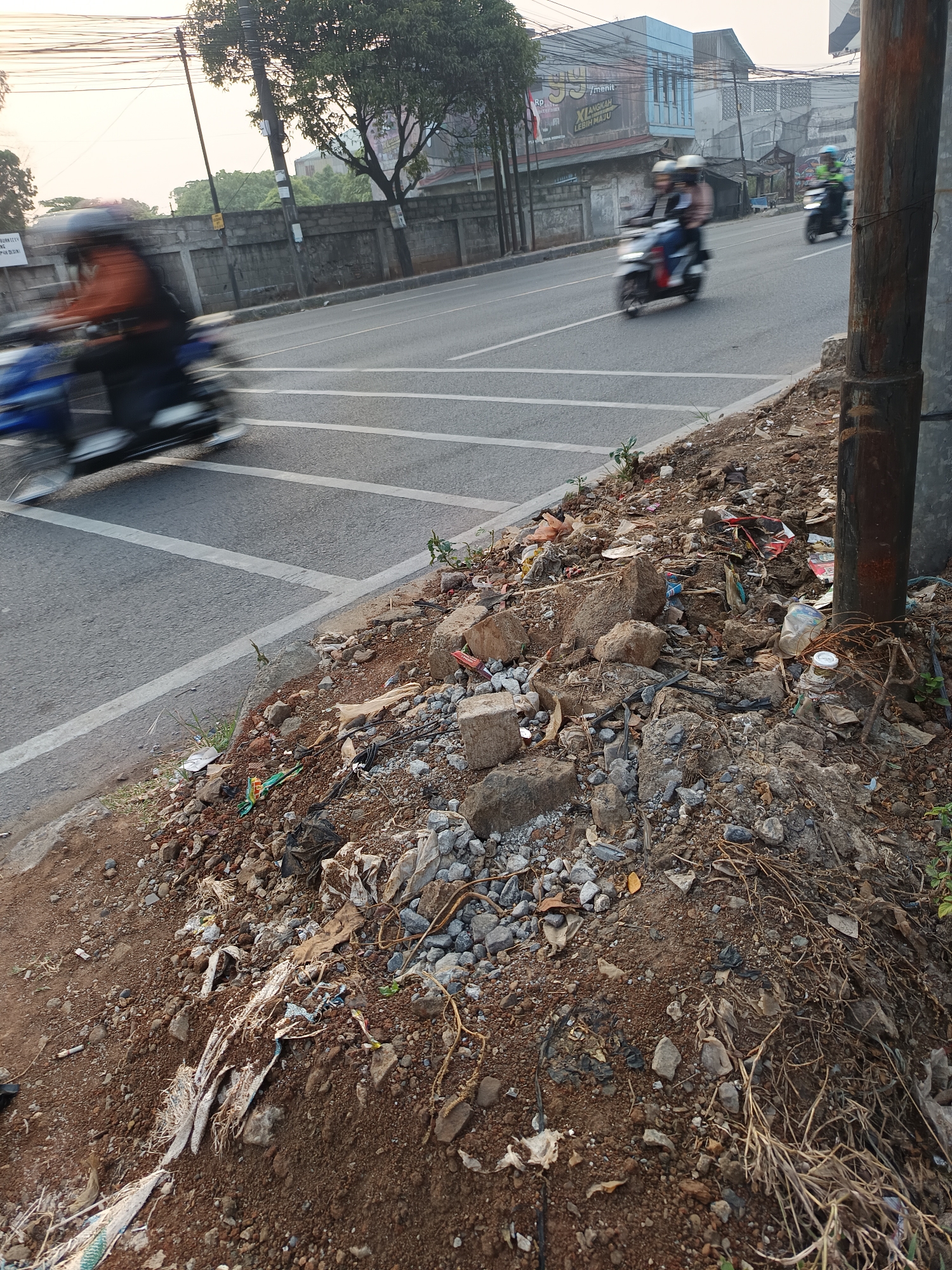 The image shows a roadside in Talaga Sari, Indonesia, littered with various types of debris, including plastic bags, bottles, and construction materials. In the background, a paved road with white lane markings is visible, with several motorcycles in motion. One motorcycle in the foreground is heavily blurred due to motion. A thick, dark pole stands on the right side of the image, partially obscuring the background. Trees and buildings can be seen further in the distance, under a bright, overcast sky. The foreground is dominated by a steep incline of dirt and rocks, heavily strewn with trash.