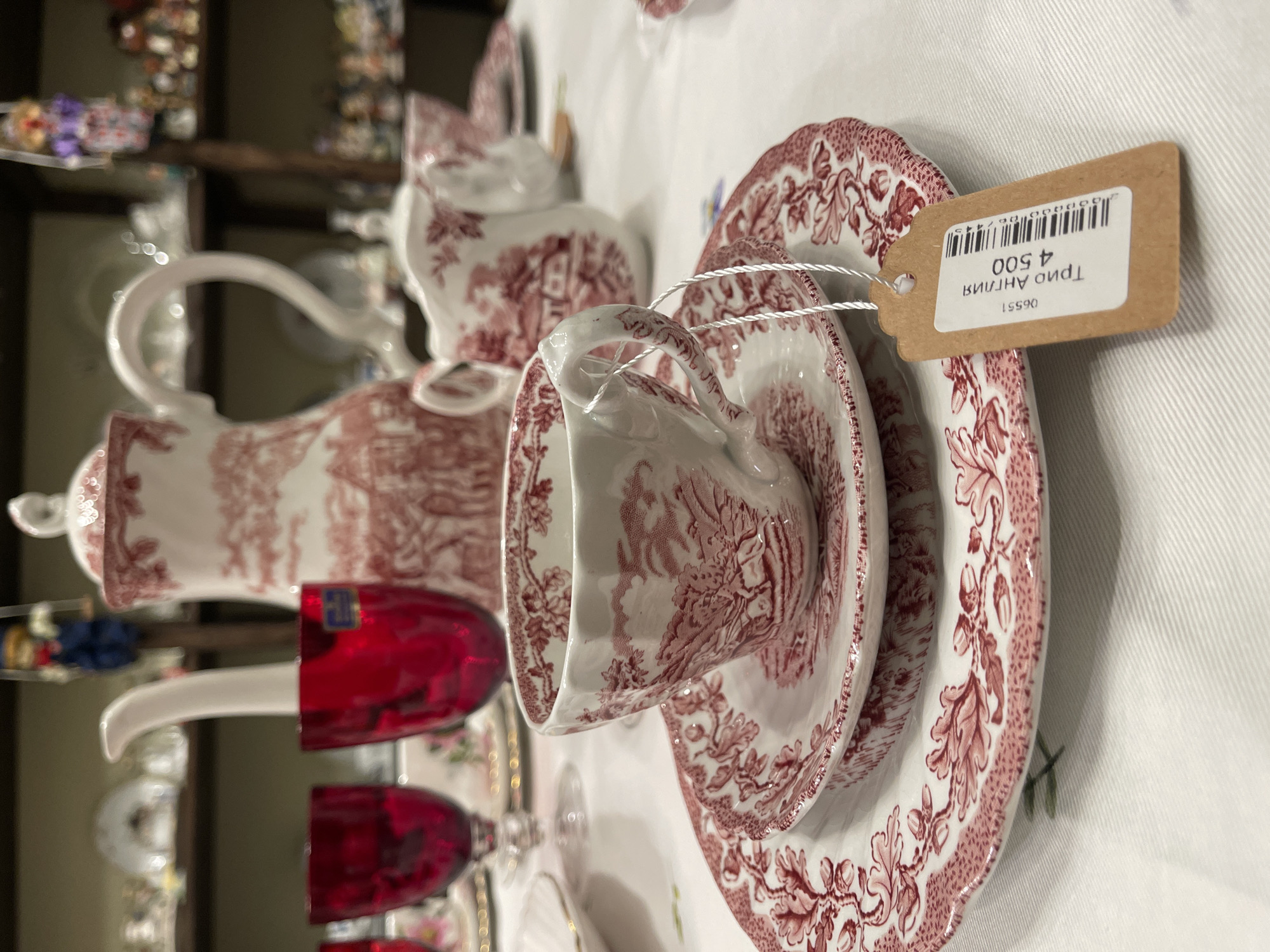 In an antique store in Svetlogorsk, Russia, a tea set with a red and white toile pattern is displayed on a white tablecloth. The set includes a teapot, a creamer, a teacup, and saucers. A price tag with Cyrillic lettering and a barcode is attached to the teacup. In the background, shelves are filled with various porcelain items and figurines. Red goblets are also visible on the table.