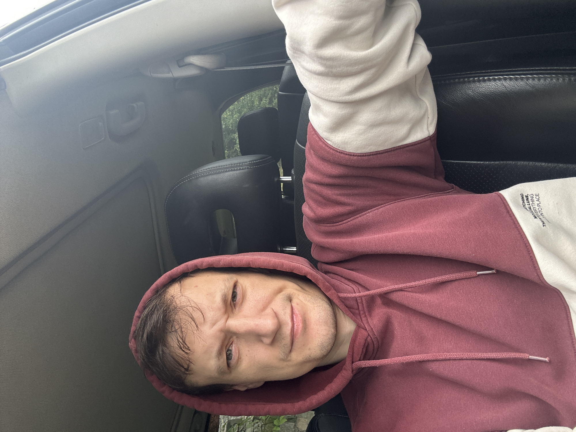 A man in a maroon and cream hoodie is sitting in the driver's seat of a car, looking at the camera. The car's interior is visible, including the dashboard, headrests, and a sunroof. The lighting suggests it is daytime, and the background visible through the windows appears to be foliage. The man has short brown hair and is clean-shaven. The image was taken in the city of Makiv, Ukraine.