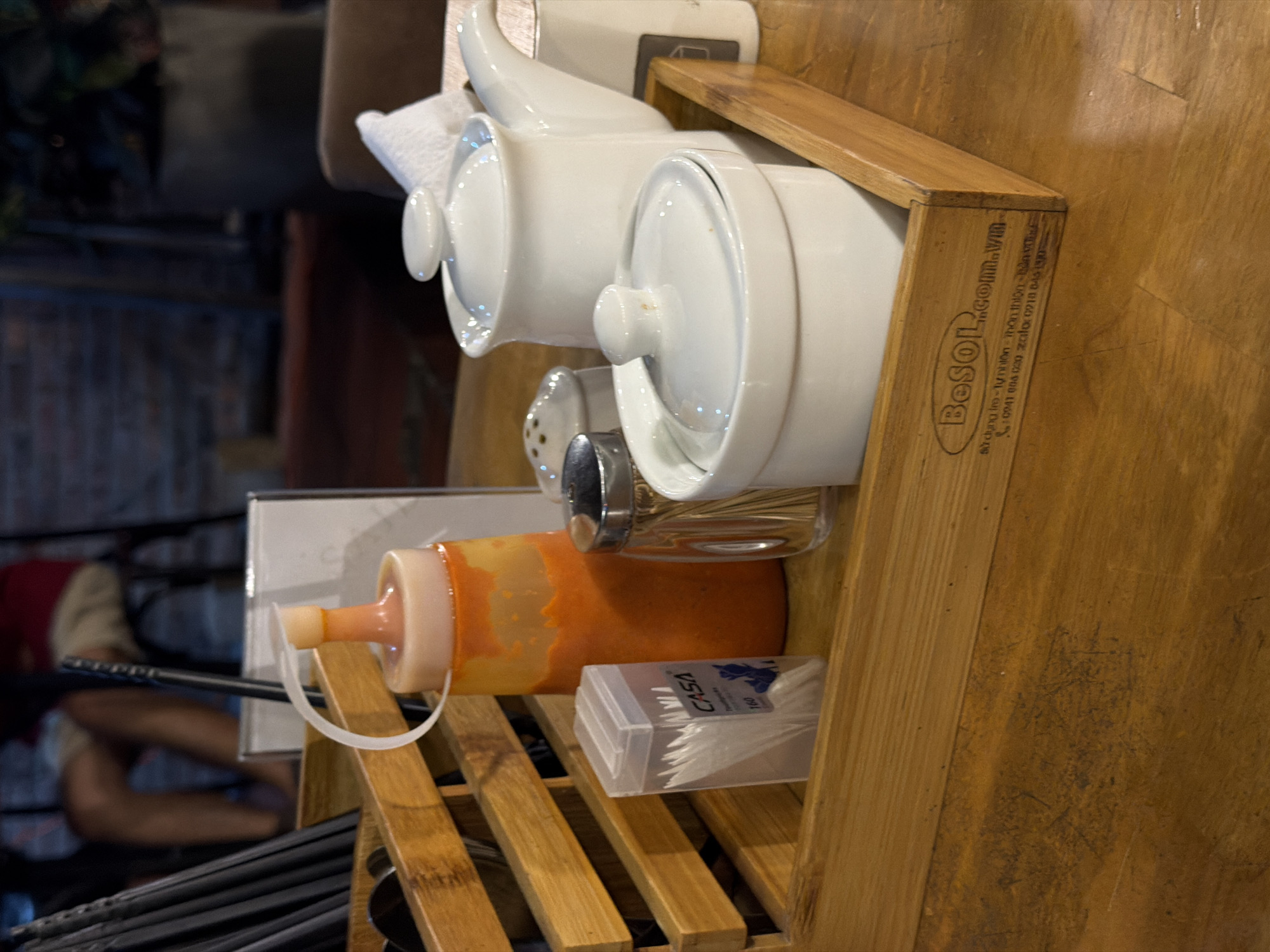 A close-up, slightly angled shot shows a wooden shelf unit, presumably in a restaurant in Đà Nẵng, Vietnam. The shelf contains various condiment containers and accessories.
On the top shelf, there are two stacked white ceramic containers with lids and a white ceramic teapot. Below these, there's a glass and stainless steel condiment dispenser, along with a plastic bottle filled with orange sauce. A box of CASA brand cotton swabs is also visible.
Beneath the condiments, the shelf holds chopsticks and spoons, with the handles pointing towards the viewer. A blurred figure can be seen in the background, near a brick wall, suggesting an occupied restaurant setting.
The wooden shelf itself has the brand name "Besol" and a website address, "Besol.com.vn", along with some Vietnamese text, likely indicating the origin and contact information of the shelf's manufacturer. The overall setting suggests a typical arrangement of condiments and utensils on a dining table or service area in a Vietnamese restaurant.