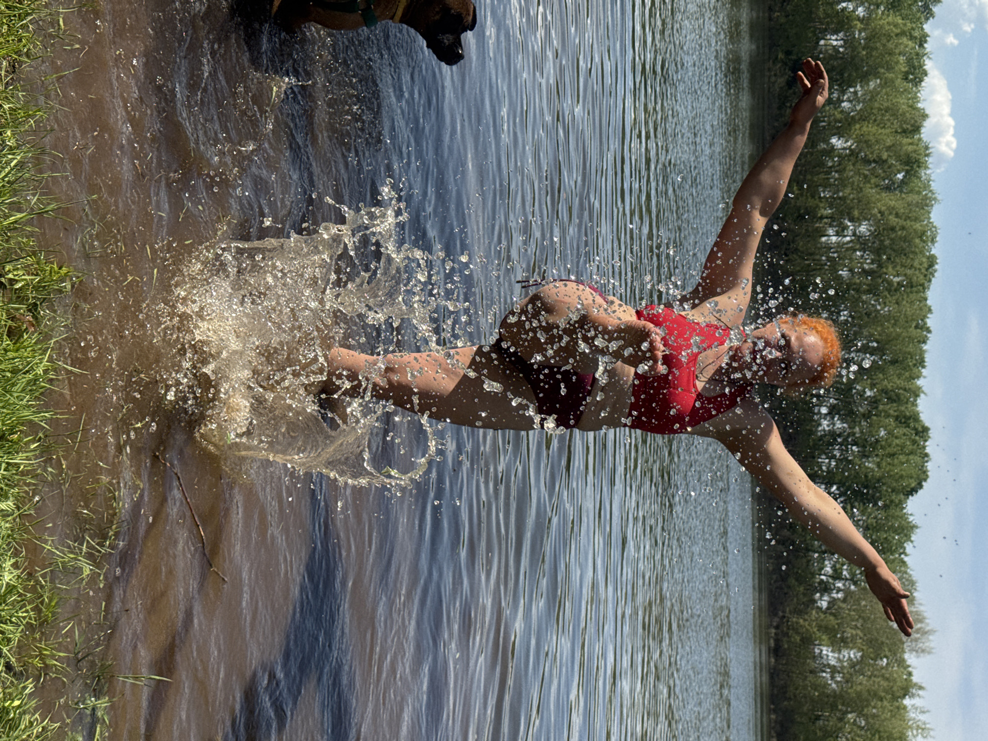 A woman with red hair wearing a red top and maroon bottoms is jumping into a body of water. Her arms are outstretched. A dog is partially visible in the upper left corner of the image. Splashes of water are visible around the woman as she enters the water. The water appears to be a lake or a wide river. Trees line the far bank. Grass is visible along the near bank. Based on the surroundings and the prompt, the location is Chaikovsky, Russia.
