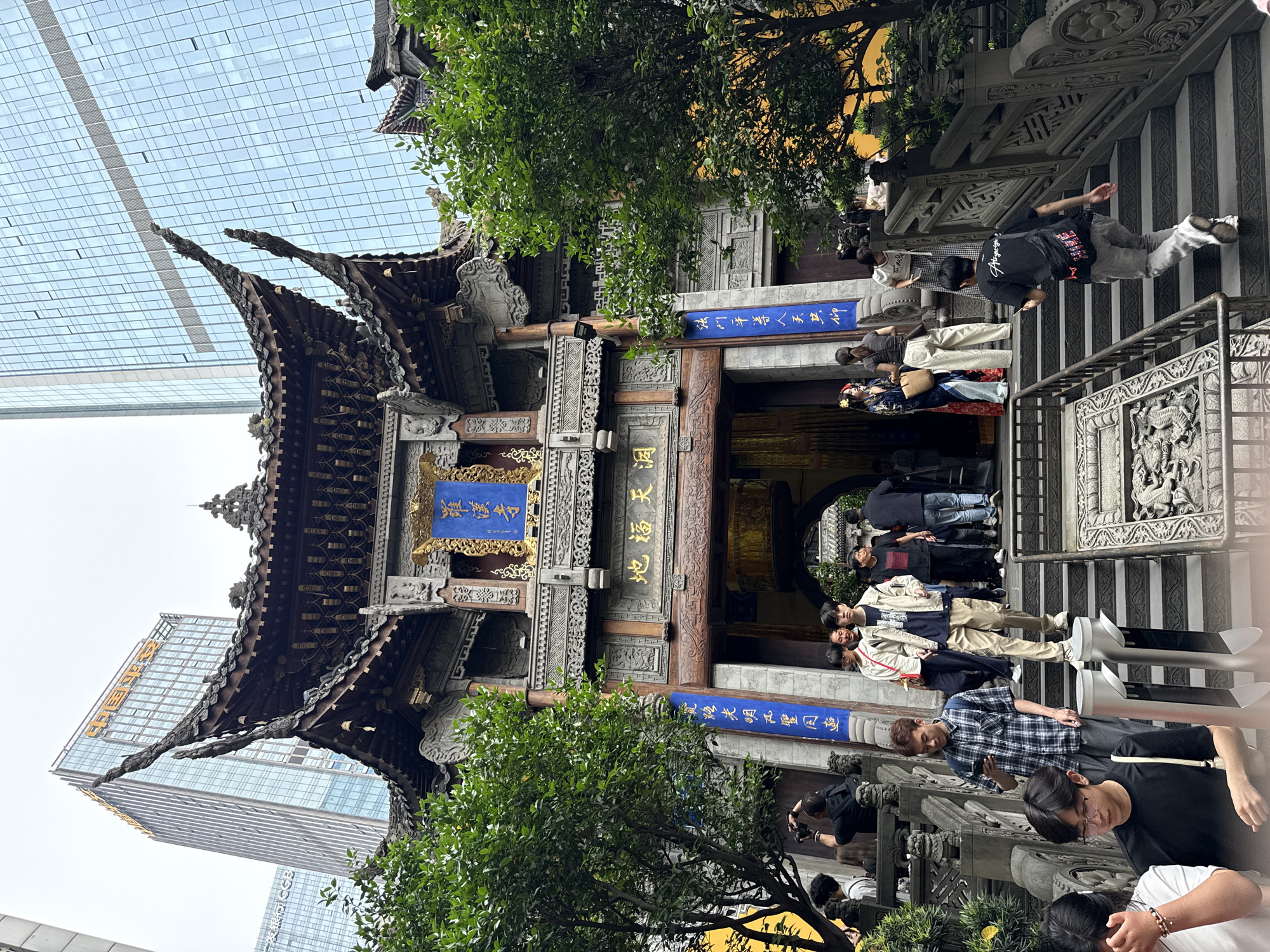 The image captures the Luohan Temple in the Yuzhong District of Chongqing, China, showcasing a blend of ancient architecture and modern urban development. The temple's traditional Chinese gatehouse is prominent, featuring intricate wood carvings, ornate roof structures, and blue plaques with golden Chinese lettering. These plaques display the temple's name and auspicious phrases like "地福天洞" (Earth's Blessing, Heavenly Cave) and "法門平導人天共仰" (The Dharma Gate Equally Revered by Humans and Gods).
Several people are visible in the foreground and around the entrance, suggesting the temple is open to visitors. Some are walking up the stairs, others are standing near the gate, and a few appear to be taking photographs. They are dressed in casual attire, reflecting a mix of ages and styles.
Juxtaposing the temple is the backdrop of modern skyscrapers, with the logos of Guangfa Bank (广发银行 GGB) and CCB (China Construction Bank) visible. This juxtaposition highlights the contrast between traditional culture and contemporary urban life in Chongqing. The lighting is diffused, indicating an overcast day, and the overall scene is captured at eye level, offering a direct and grounded perspective. Trees and foliage add a touch of nature, softening the contrast between the temple and the surrounding buildings.
