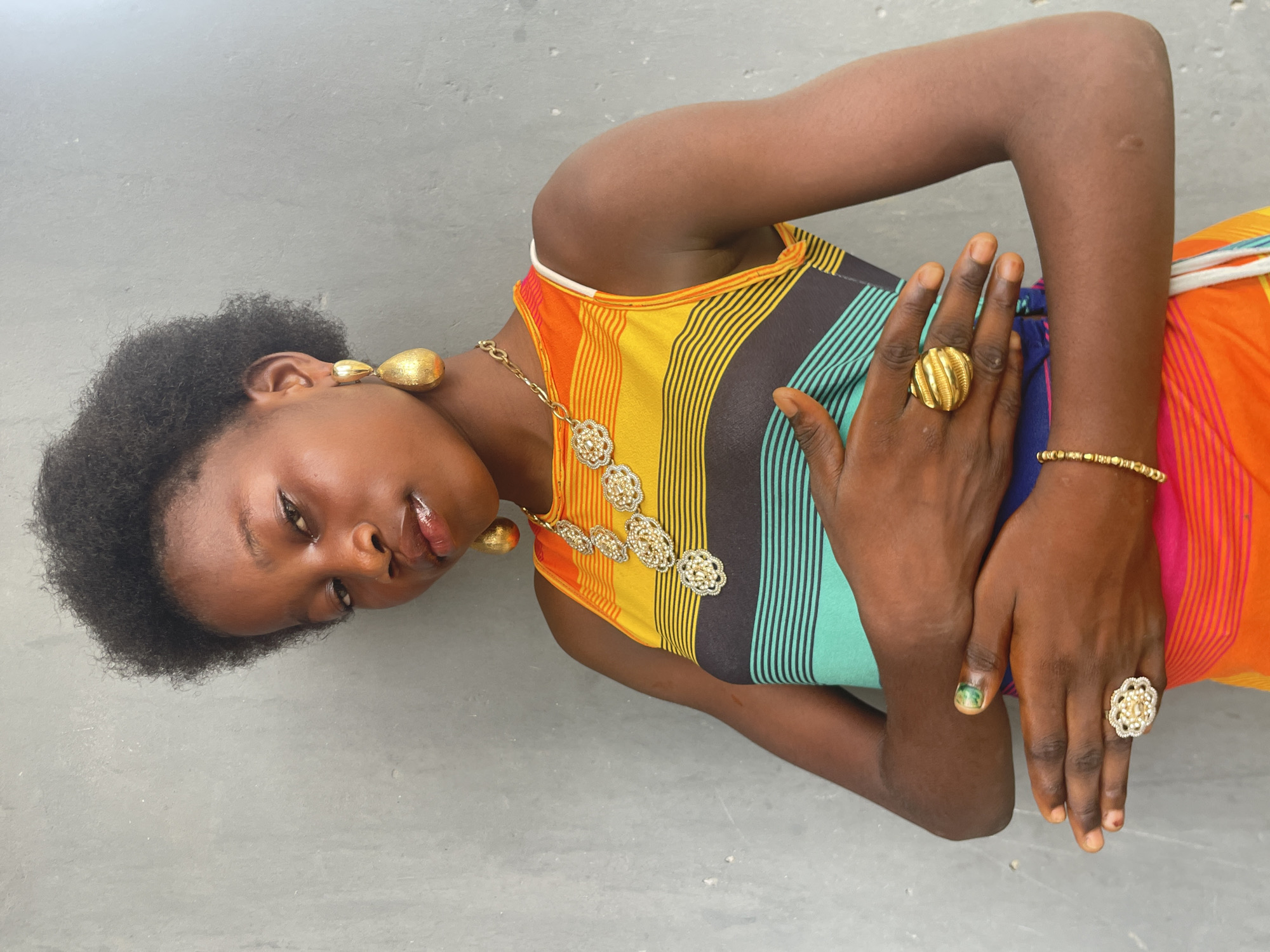 A young woman with dark skin and short, curly dark hair is depicted lying down or reclining against a plain, light grey background, likely a concrete floor or wall. The image is taken in Badagry, Nigeria. She looks directly at the camera with a calm and direct expression, her lips slightly parted and glossy.
She is wearing a sleeveless top or dress characterized by vibrant horizontal stripes in colors such as orange, yellow, black, and teal. Her attire is complemented by elaborate gold-toned jewelry. She wears large, tear-drop shaped earrings, and a prominent necklace adorned with several intricate, round, floral-patterned medallions. Her right wrist features a delicate gold beaded bracelet, while her left index finger bears a large, textured gold ring. Another decorative ring with a floral design is visible on her right ring finger. Her fingernails are short and natural, with a hint of green polish remaining on her left thumbnail. The lighting is bright and diffused, suggesting daytime. There is no visible text in the image.