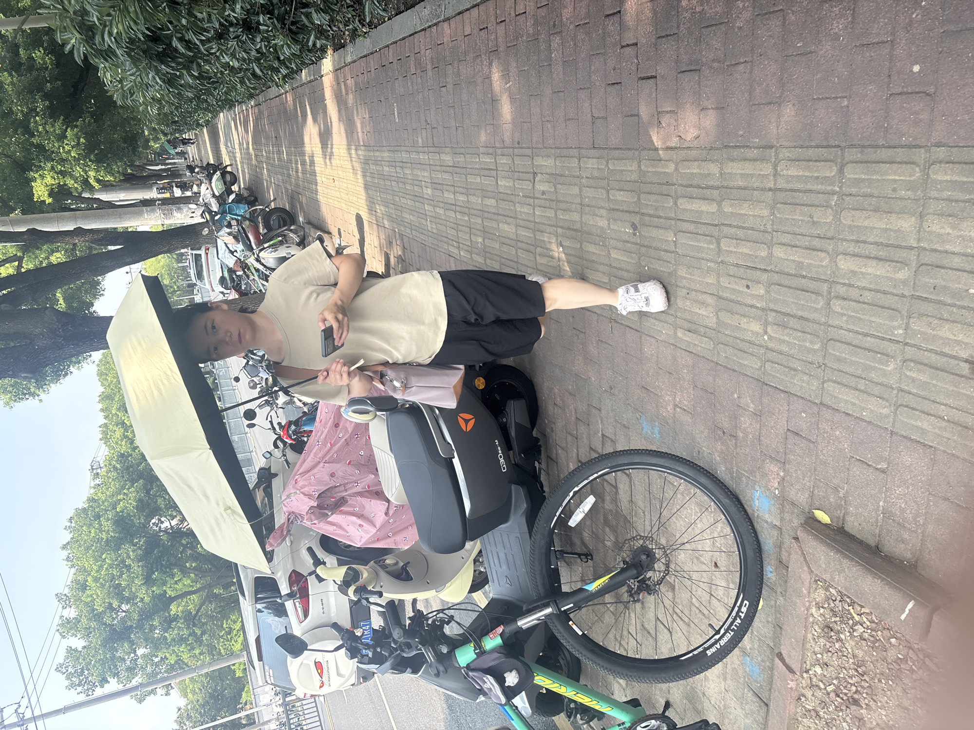 A young woman walks on a sidewalk holding a light-colored umbrella over her head. She is wearing a tan t-shirt and black shorts, white sneakers, and a brown shoulder bag. She is looking down at her mobile phone, which she holds in her right hand. The sidewalk is paved with brick and has tactile paving for the visually impaired. In the background, several parked motorcycles and scooters are visible, along with a white SUV. A green bicycle is parked in the foreground to the left of the woman. The scene is located on a tree-lined street in Suzhou Industrial Park, China, under a bright, sunny sky.