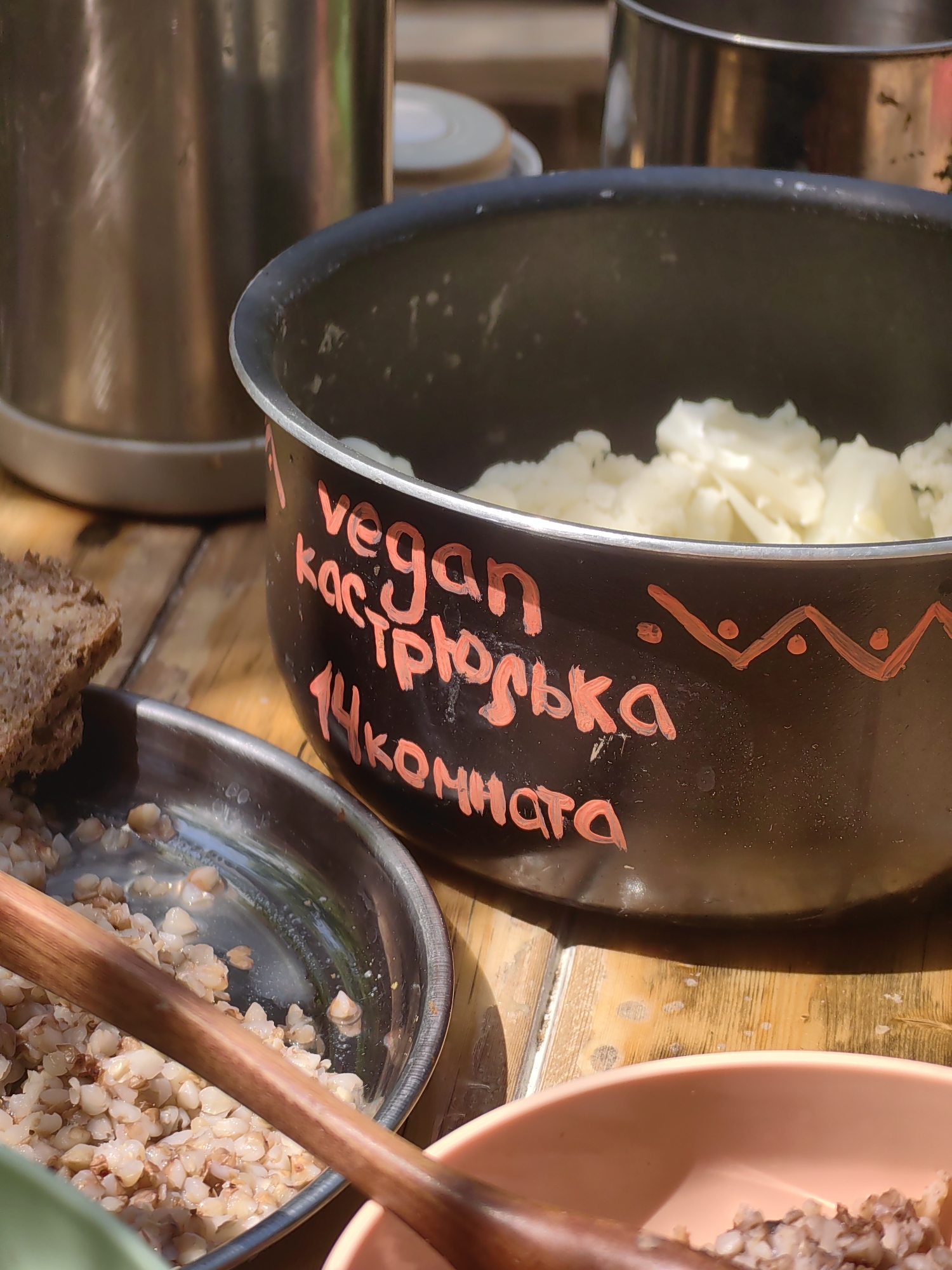 A close-up, eye-level shot shows a serving of cooked buckwheat in a metallic bowl, a slice of dark bread, and a black pot with a white filling, possibly mashed potatoes or cauliflower. The pot has "vegan кастрюлька 14 комната" written in red paint on its side. The scene appears to be outdoors, with sunlight illuminating the food and a wooden surface beneath the bowls.