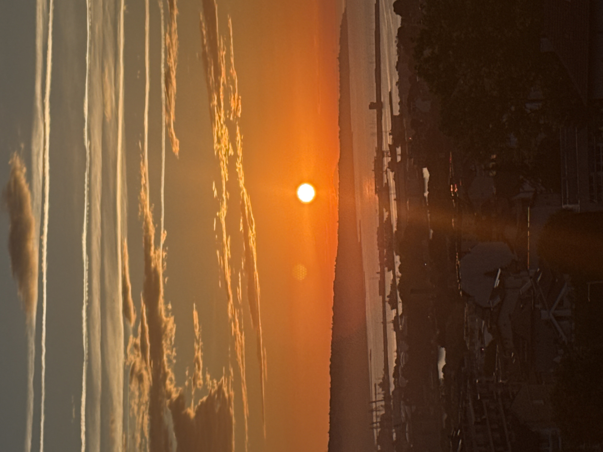 An aerial, high-angle, vertical shot captures a vibrant sunset over a coastal city. The sun, a bright orange orb, is positioned slightly left of center in the upper half of the image, casting a warm glow across the sky and water. Multiple layers of clouds, some thin and white like contrails, stretch horizontally across the sky, intermingled with wispy orange and yellow clouds.
Below the sky, a silhouetted hilly coastline is visible, bordering a body of water that reflects the sunset's colors. In the middle ground, a port or harbor area is dotted with numerous ships of varying sizes, cranes, and structures. The foreground is a densely packed cityscape with buildings and trees, all rendered in dark silhouettes due to the low light. The overall impression is of a serene evening over an active port city.