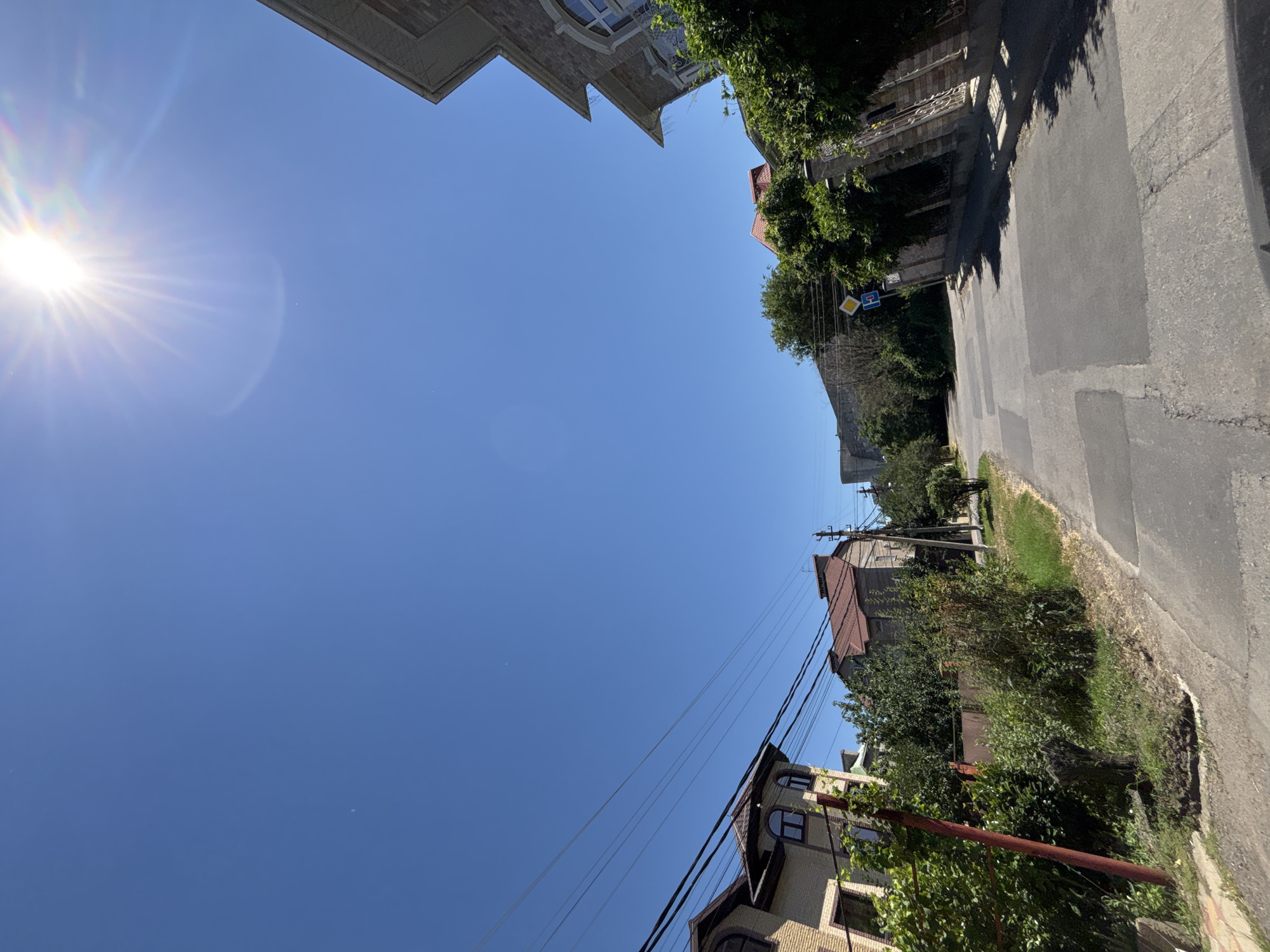 The image captures a sunny day in Krasnodar, Russia. The scene is dominated by a bright blue sky and the sun, which is visible in the upper left quadrant, casting a lens flare and rays of light. The perspective is tilted, with the ground and buildings appearing at an angle.
The foreground shows a paved road running diagonally across the right side of the image. Along the roadside, there is a strip of green grass and dense vegetation. Several houses with tiled roofs are visible, interspersed with trees. One house in the lower right has a light-colored brick facade and multiple windows, some with arched tops. Another house, further up the road, has a darker facade and a red tiled roof. Power lines crisscross the sky above the houses.
In the upper portions of the image, the rooftops of buildings are visible, suggesting an urban or suburban setting. One building on the far left appears to be a multi-story structure with ornate architectural details, including arched windows. Lush green foliage spills over the edge of another building on the right, adding to the sense of a warm, summery environment. Two road signs are visible on poles near the road, though their text is not clearly legible. The overall impression is one of a bright, clear day in a residential area, with a strong emphasis on the natural light and the sky.