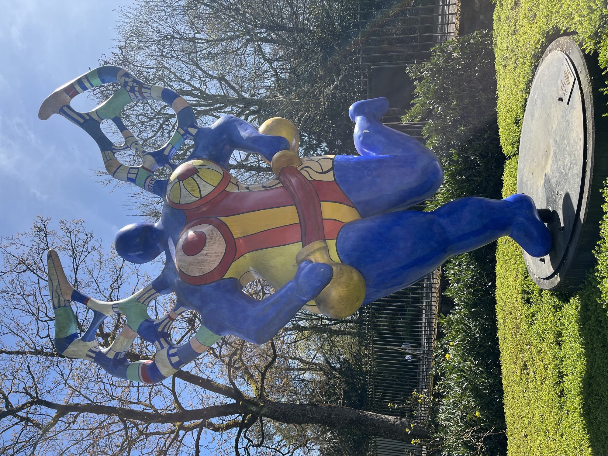 The image features a colorful, female sculpture by Niki de Saint Phalle, known as a "Nana", located in the municipal park of Luxembourg City, Luxembourg. The sculpture is predominantly blue with yellow and red accents, depicting a stylized female figure with raised arms holding yellow balls. The figure is set on a circular metal base, surrounded by a trimmed hedge. In the background are trees with bare branches against a blue sky, and a metal fence. The image is taken during daylight with natural lighting.