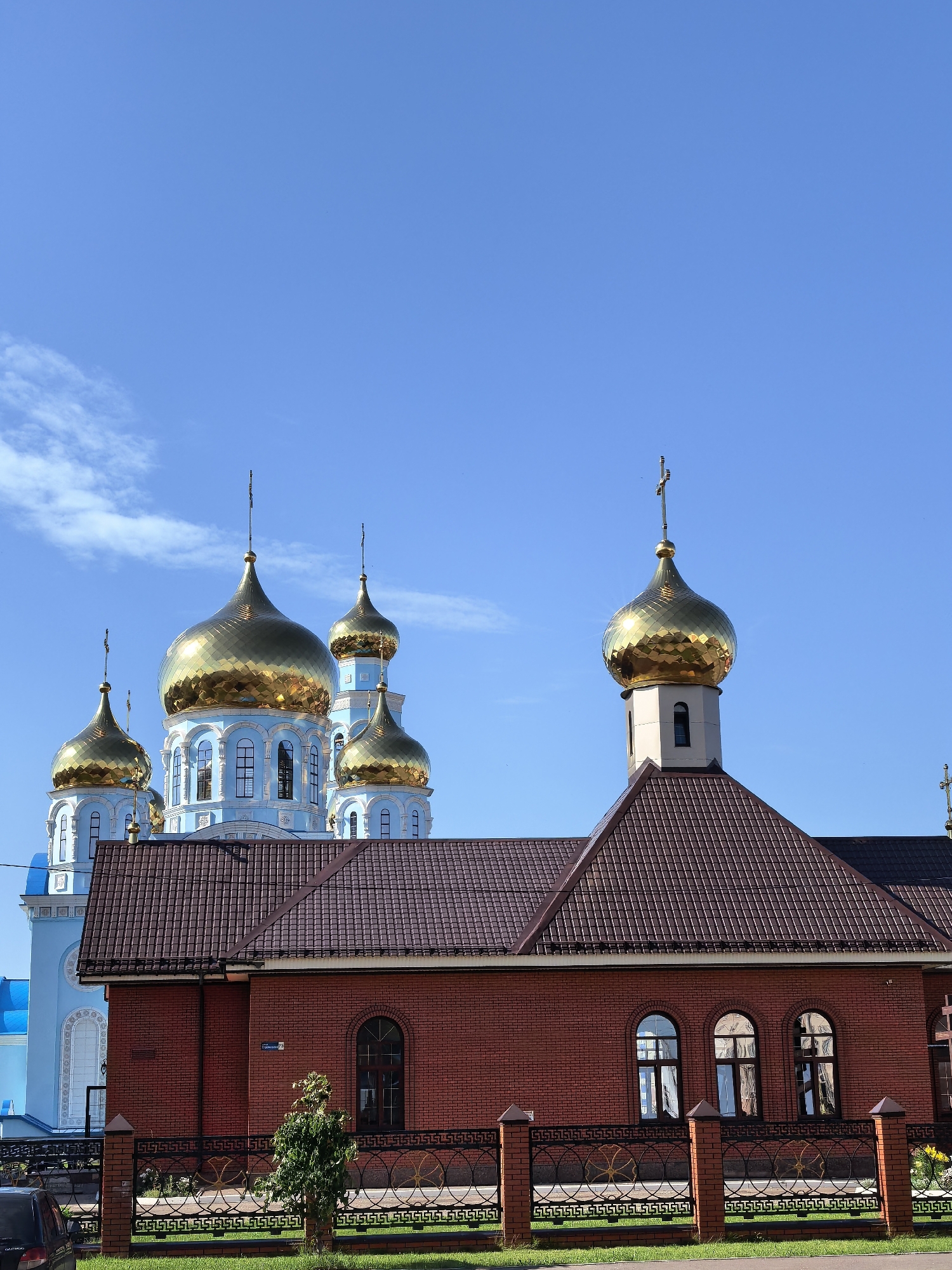 The image shows a bright, sunny day with a clear blue sky. In the foreground, a red brick building with a brown tiled roof is visible. This building has arched windows and a decorative fence in front of it. Behind this building, a larger, light blue structure with multiple golden domes can be seen. These domes are ornate and reflect the sunlight. The scene appears to be a religious complex, likely a church, given the presence of golden domes topped with crosses. The location is identified as the Church of the Resurrection of Christ in Sterlitamak, Russia. There are no people or specific actions depicted in the image.