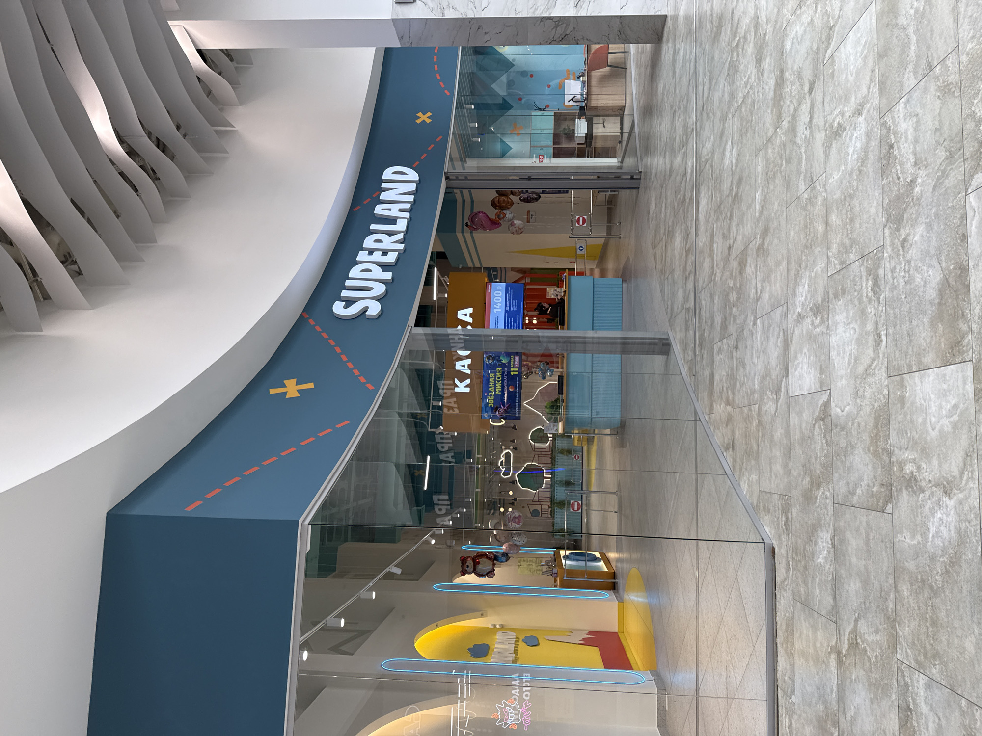 The image captures the entrance to "Superland," a children's entertainment center, likely located in a shopping mall in Izhevsk, Russia. The entrance is defined by a curving blue wall with the "SUPERLAND" logo in white, with a yellow X and dotted orange lines evoking a map or pathway. The facade is mostly glass, offering a view into the play area, which includes a yellow and red display with blue neon accents and various decorations and advertisements. Beyond the glass, a reception area with a blue counter and staff can be seen. The flooring is a neutral-toned tile.
