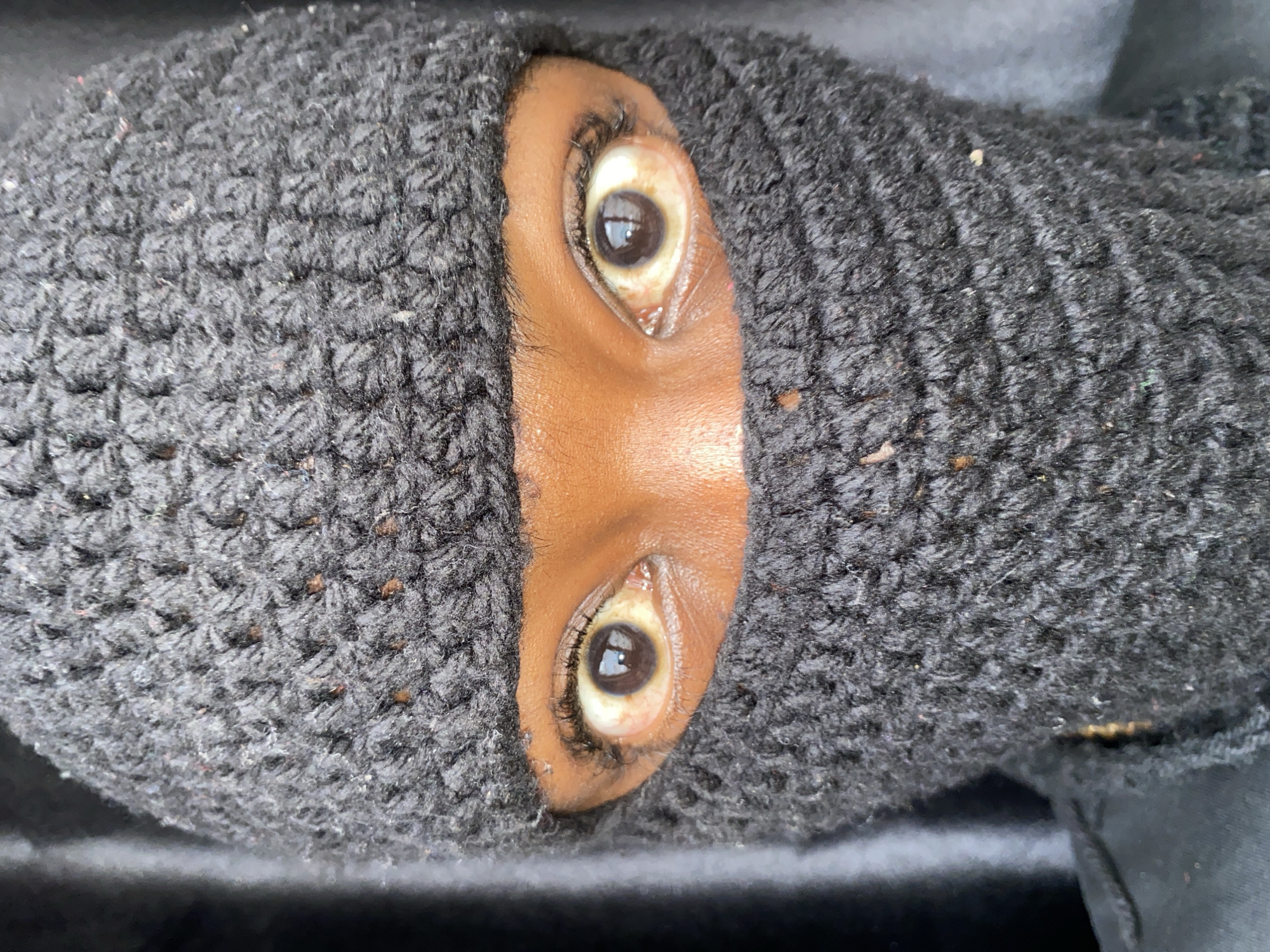 A close-up photograph shows a person wearing a black knit balaclava. Only the eyes and a small portion of the person's nose and forehead are visible through an opening in the balaclava. The person has brown skin and dark eyelashes. The balaclava appears to be made of a thick, textured yarn. The background is out of focus and dark. The specific location in Barakin Akawo, Nigeria cannot be determined from the image.