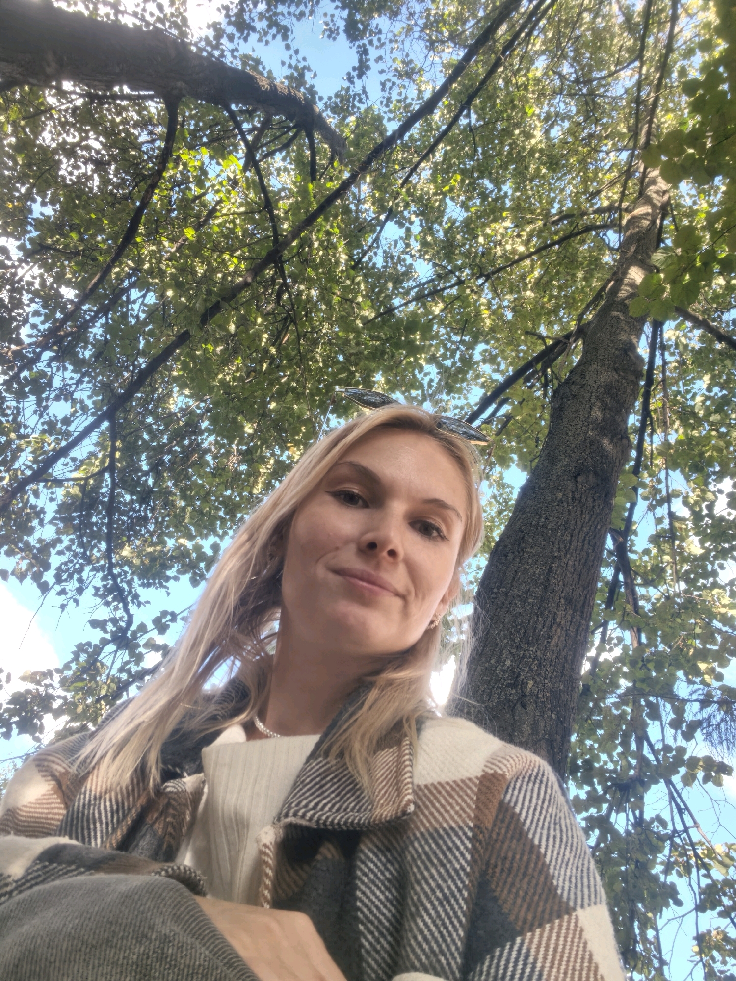 A young woman with blonde hair, wearing a plaid jacket and a pearl necklace, is looking up at the camera from beneath a canopy of green leaves and tree branches against a bright blue sky. She is positioned at the bottom of the frame, with a thick tree trunk to her right and branches extending across the top. The scene suggests an outdoor setting, possibly a park or a street lined with trees, in Sterlitamak, Russia.