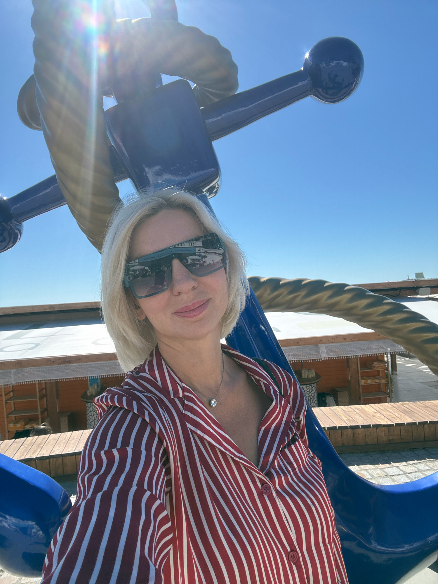 The image shows a blonde woman in sunglasses and a red and white striped shirt taking a selfie outdoors. She is standing in front of a large blue and gold anchor sculpture against a clear blue sky. In the background, there are some wooden structures with white canopies, possibly part of a beach resort or cafe. The scene appears to be in a sunny, outdoor urban environment, consistent with a location in Sochi, Russia.