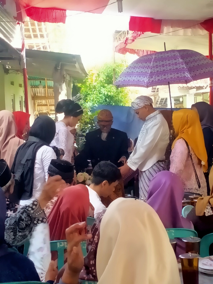 A wedding ceremony unfolds outdoors, shaded by red and white awnings. The bride in white, adorned with a tiara, stands beside the groom. A man in a suit oversees the event, with another assisting the couple, holding their hands. A purple umbrella provides additional shade. Guests, many wearing hijabs, fill the space. Tables are set with glasses and plates, ready for the reception.