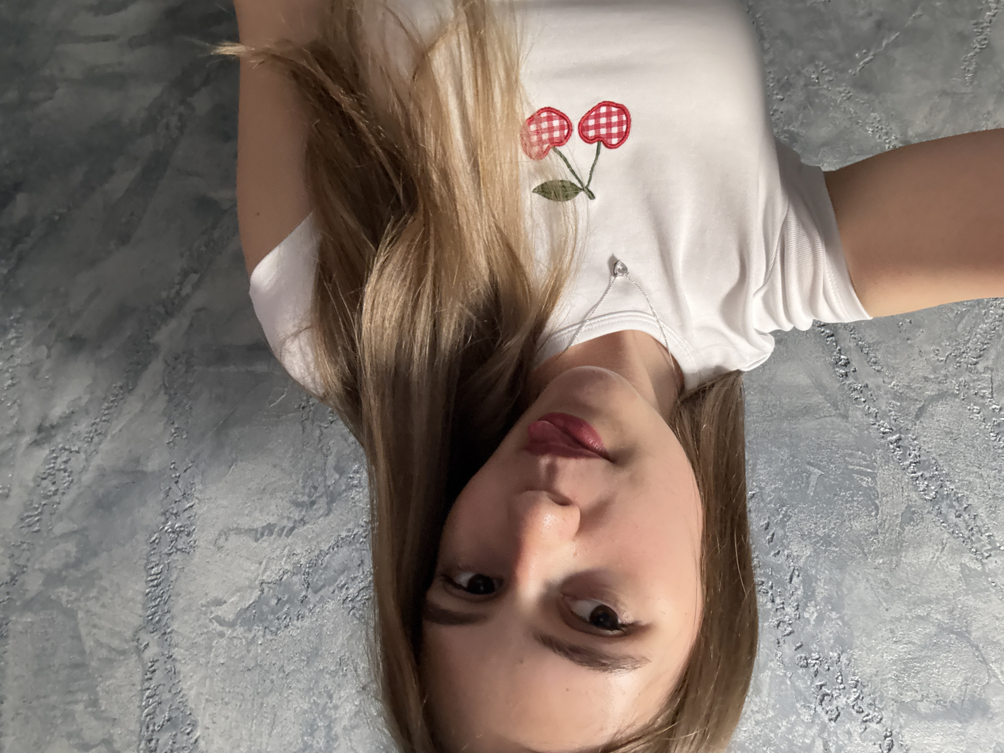 A young woman is seen in a tilted close-up shot. She is wearing a white t-shirt with a cherry design on the front and a thin silver necklace. The background is a textured wall painted in shades of grey and blue. The location, Panagyurishte, Bulgaria, cannot be confirmed based solely on the image.