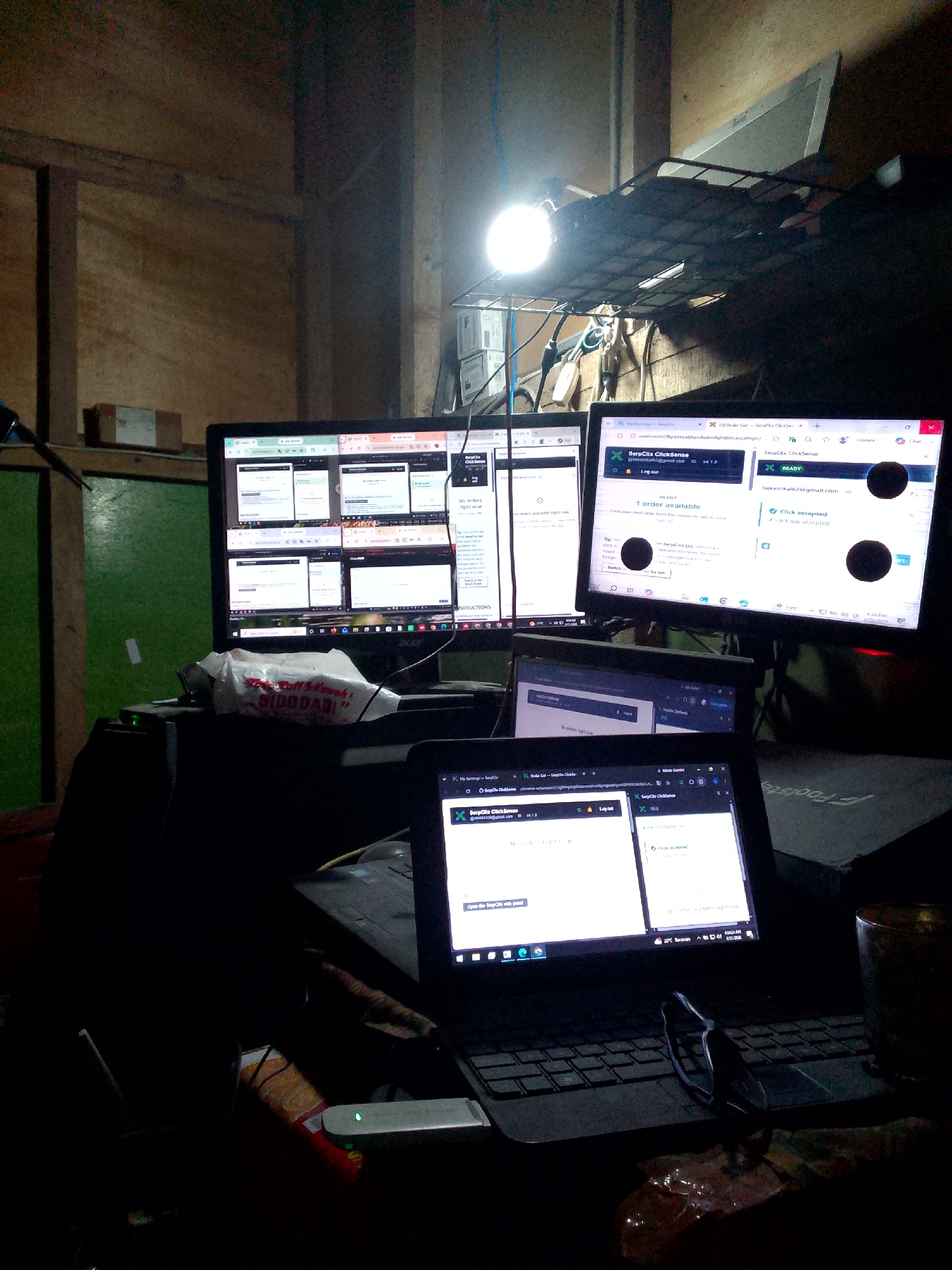 The image depicts a dimly lit indoor workspace, likely a home office, with multiple computer monitors and a laptop actively displaying web pages. The environment appears to be constructed of rough, unfinished wood panels, suggesting a rustic or improvised setting. A bright overhead lamp illuminates a metal shelf unit positioned above the monitors.
On the primary large monitor, several browser windows are open, showing what appears to be a web application with lists and data. The right side of this monitor displays a website titled "SerpClicks ClickSense" with sections indicating "READY" and "1 order available," along with user information and a "Log out" button.
A laptop is positioned in the foreground, also displaying "SerpClicks ClickSense" in its browser window. A pair of glasses rests on the laptop keyboard. In front of the monitors, a plastic bag containing what looks like food items is visible. A USB modem with a green indicator light is situated on a dark surface to the left of the laptop.
The overall impression is of a functional but unpolished workspace, possibly used for online work or tasks. The presence of multiple screens and the active web applications suggest sustained computer usage. The lighting is functional but creates strong shadows, contributing to the somewhat cluttered and utilitarian atmosphere. The location is identified as Gunungmasigit, Indonesia.
