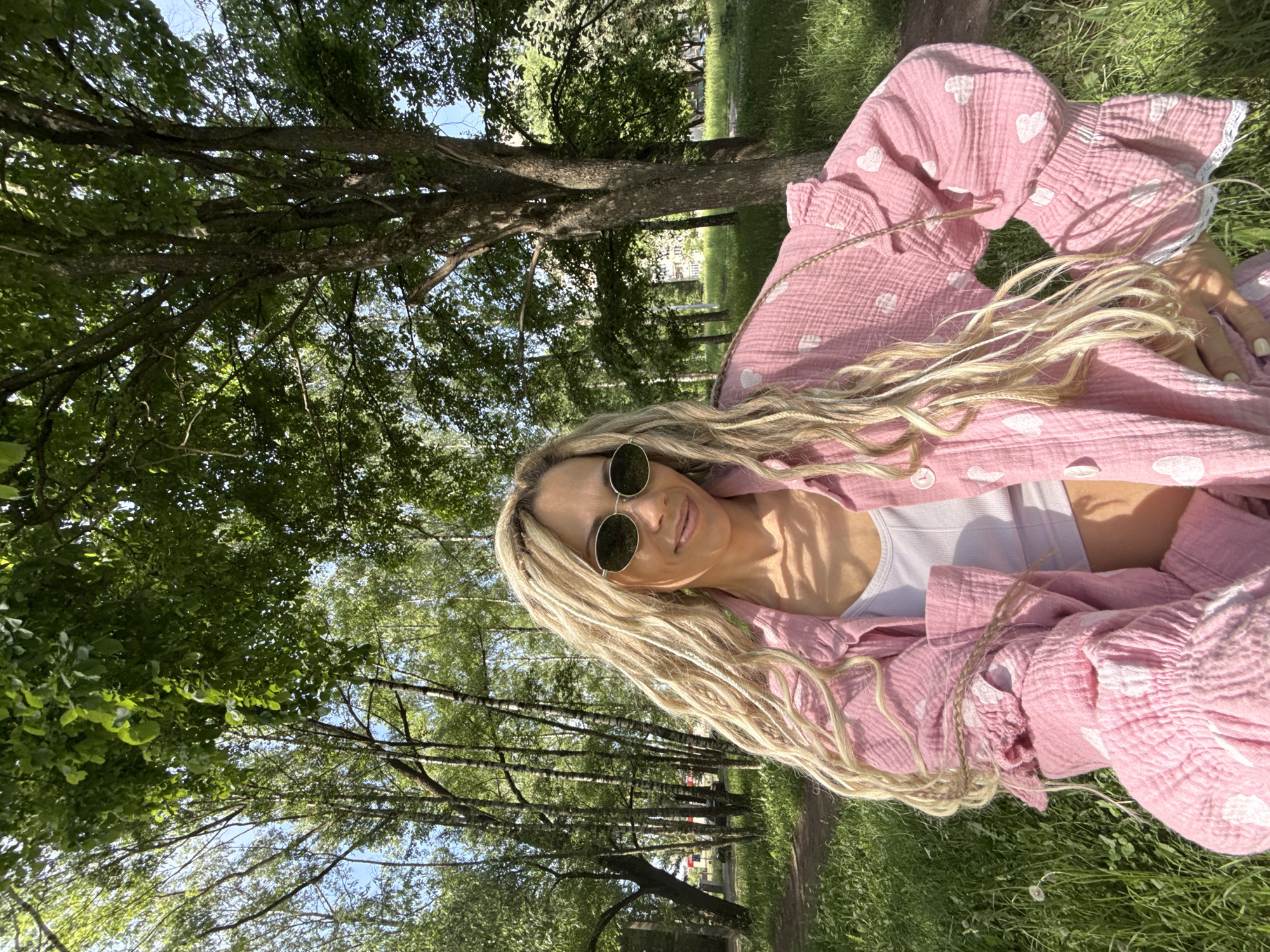 A woman is depicted in a park or wooded area within the city of Rusanovo, Russia. She has long, blonde, wavy hair styled with visible dreadlocks or braids and wears dark, oval-shaped sunglasses. She is smiling and looking directly at the camera. Her attire includes a light pink, textured fabric shirt with white heart patterns and ruffled, lace-trimmed cuffs, worn open over a white crop top. Her right hand is positioned on her hip. The background features a dense canopy of green deciduous trees, including slender birch trees on the left and a larger, darker-barked tree on the right. Lush green grass covers the ground, with a visible dirt path winding through it on the left. Patches of clear blue sky are visible through the foliage, and a faint urban structure or building is discernible in the distant background through the trees on the right. The scene is illuminated by natural sunlight, creating shadows and highlights.