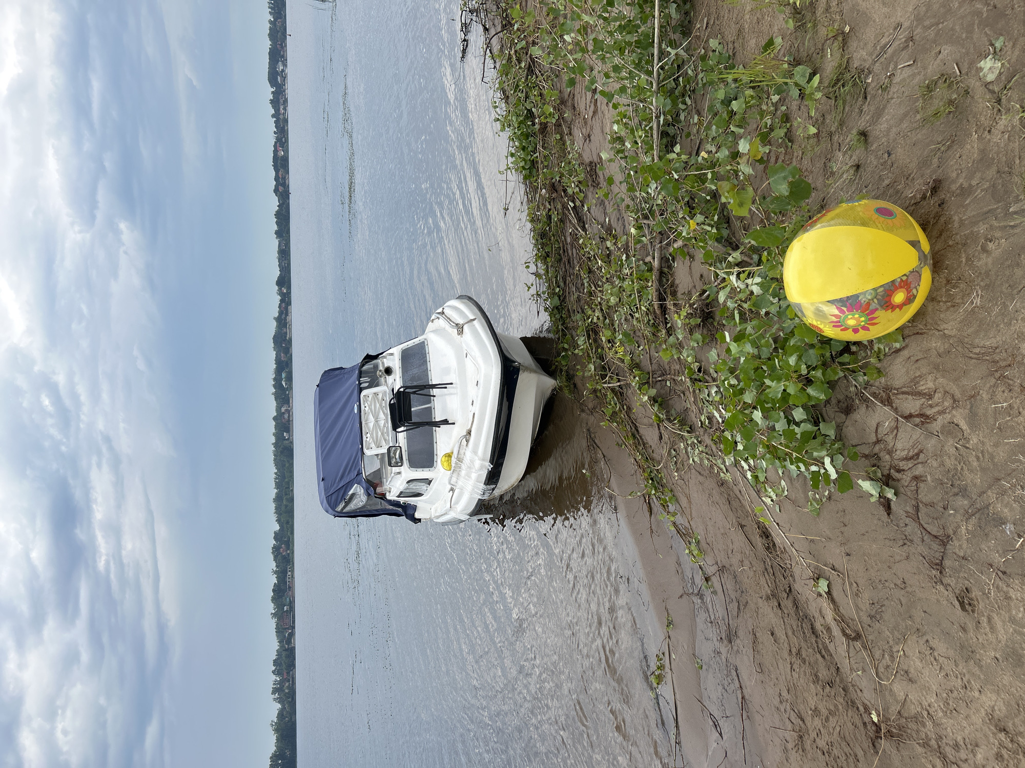A white boat with a blue canopy is docked on the muddy shore of a wide river. The boat has a black stripe along its waterline and a white railing. In front of the boat, on the sandy bank, lies a deflated yellow beach ball with colorful floral patterns. The riverbank is covered in debris, including branches and green foliage. In the distance, across the water, a treeline with houses is visible under a cloudy sky. The location is the riverbank of Novokuybyshevsk, Russia.