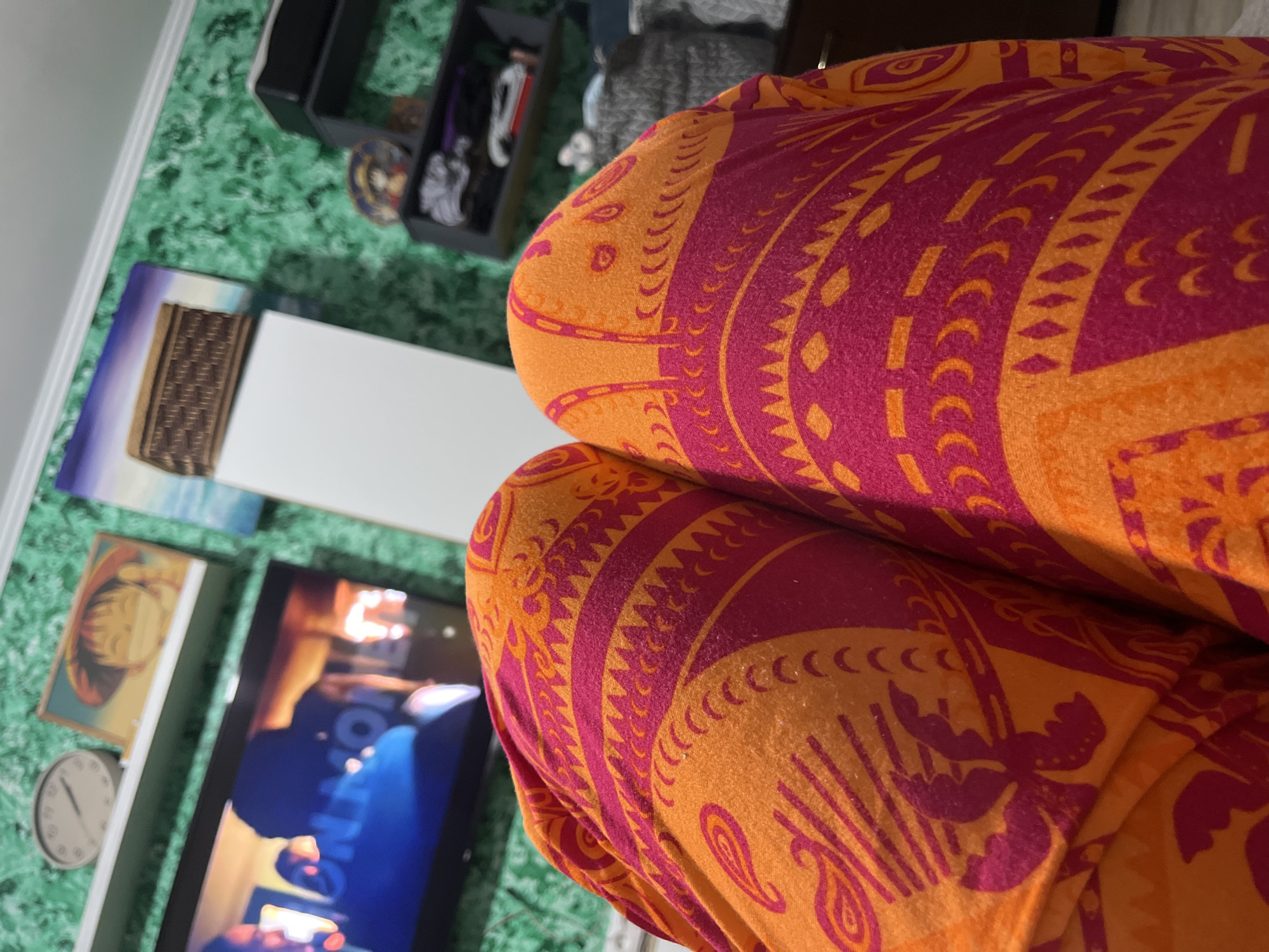 The image shows a close-up view of a person's legs covered in orange and pink patterned pajamas, likely in a home environment. In the background, a television screen displays a blurry image with text, a white shelf with a clock and framed picture, a textured green wall, and dark shelves with various items. It is impossible to determine the exact location in Сеніцкі сельскі Савет, Belarus, or identify any specific people or actions from this image.