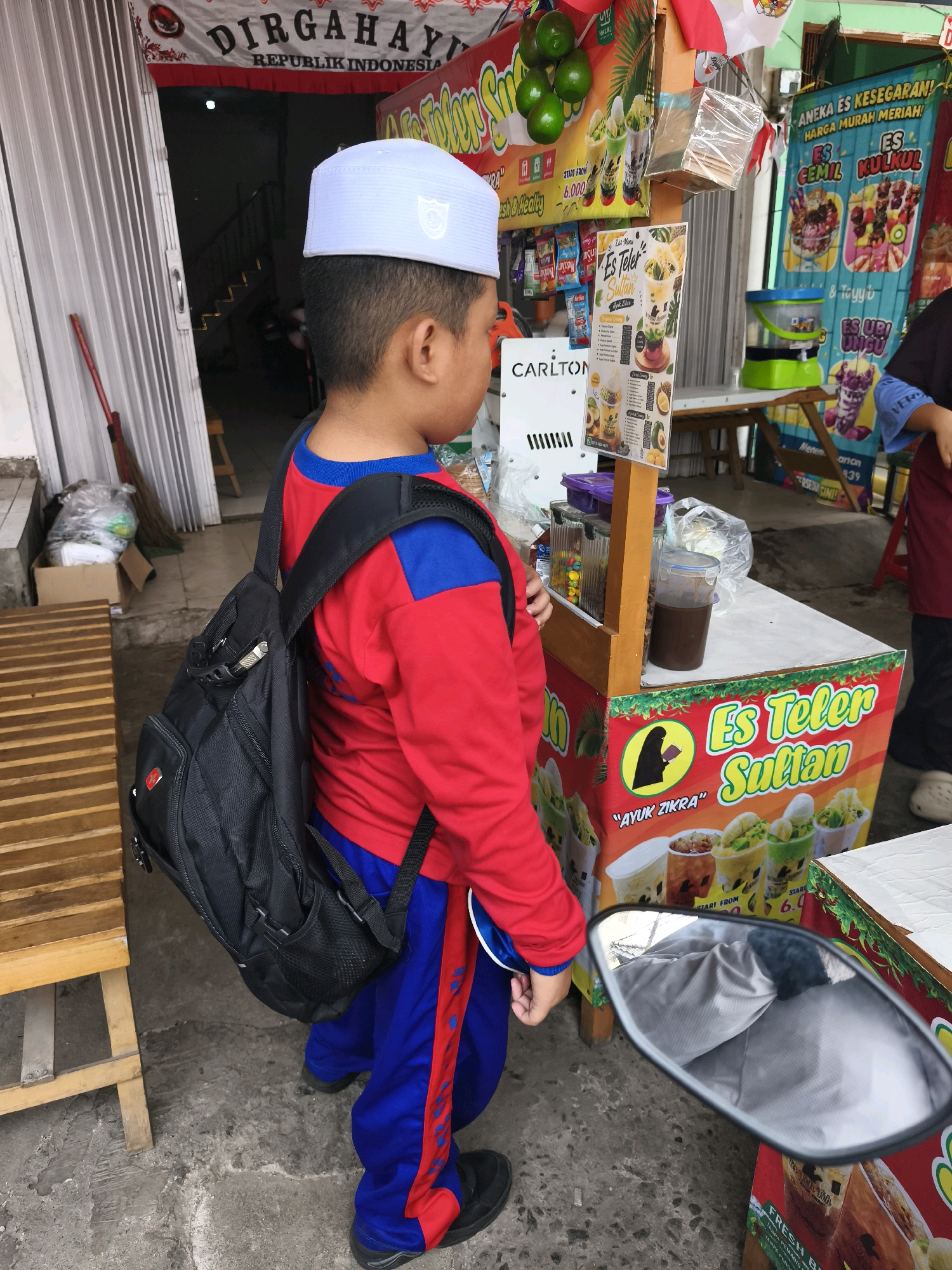 A young boy wearing a white cap and a red and blue shirt stands next to an outdoor food stall in Palembang, Indonesia. He carries a black backpack. The food stall is decorated with colorful banners advertising various drinks and desserts, including "Es Teler Sultan" and "Es Kulkul." Several bottles of drinks and jars of ingredients are visible on the counter. In the background, a metal gate is partially open, revealing a staircase. The ground is paved with concrete. A motorcycle mirror reflects a gray surface.