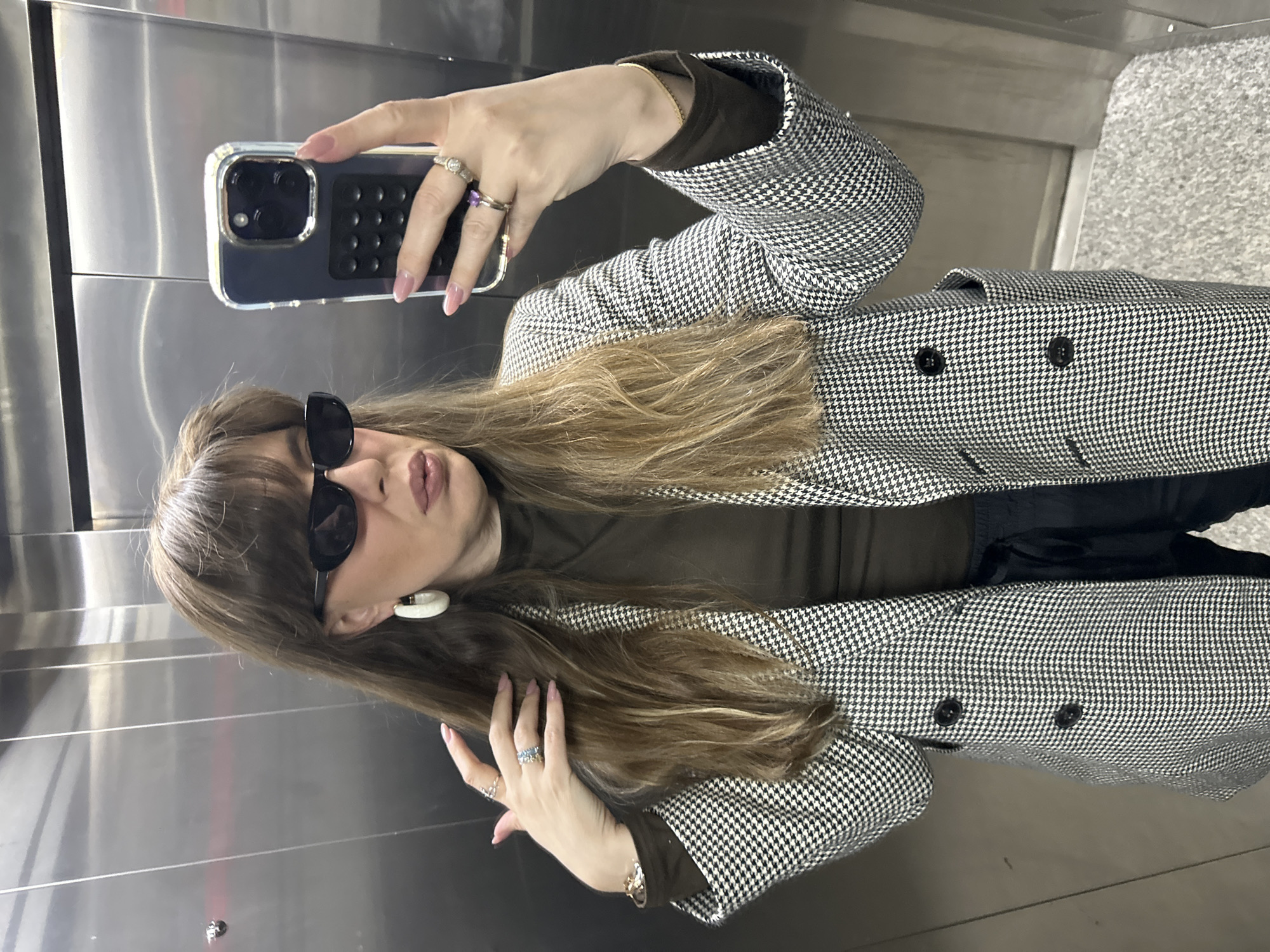 A woman stands inside an elevator, taking a selfie with her smartphone. She has long, light brown hair with bangs, and she's wearing dark sunglasses and a large white hoop earring. She's dressed in a brown turtleneck and a black and white houndstooth blazer. Her nails are painted light pink, and she wears several rings on her fingers. The interior of the elevator is metallic, likely stainless steel, and there is a concrete floor visible. There is no indication this image was taken in Tbilisi, Georgia.