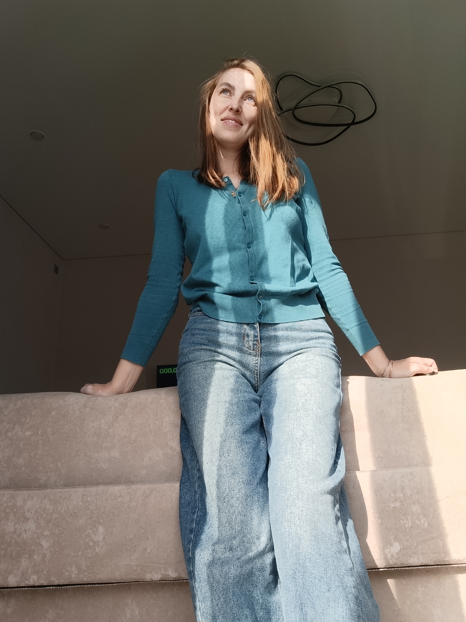 A young woman stands in a brightly lit, indoor space, leaning against a stack of what appears to be upholstered furniture. She is wearing a teal cardigan and light blue jeans. Her gaze is directed upwards and to the left, and she has a slight smile. The lighting suggests it is daytime. In the background is a dark, indeterminate area, possibly the interior of a room with a modern, abstract ceiling light fixture. A small digital display emitting a green light can be seen at the edge of the furniture. There is no visual evidence in the image to confirm the specific location of Chaikovsky, Russia.