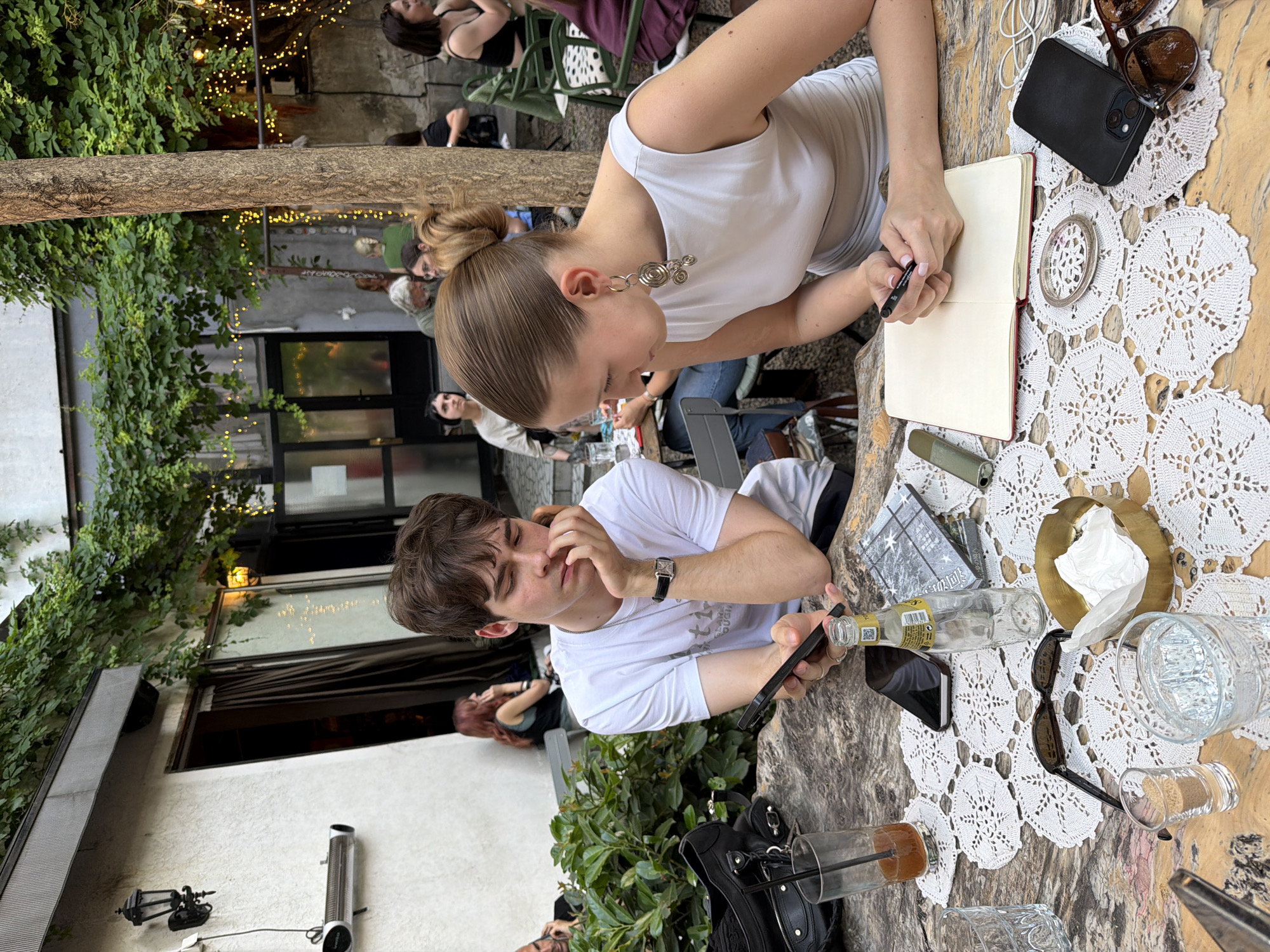 The image captures a casual outdoor scene, possibly a cafe or restaurant in Belgrade, Serbia. Two individuals are central to the frame: a woman with her hair pulled back, wearing a white top, is engrossed in writing in a notebook with a pen. Adjacent to her, a man in a white t-shirt holds a mobile phone. The table between them is covered with a lace tablecloth and holds several items, including sunglasses, empty bottles, drinking glasses, a metal trinket, and a black mobile phone. Other people are visible in the background, seated at tables. The outdoor setting is embellished with greenery, including climbing plants on the building facade and trees overhead, adorned with string lights. The overall ambiance suggests a relaxed, social gathering in a lively urban environment.