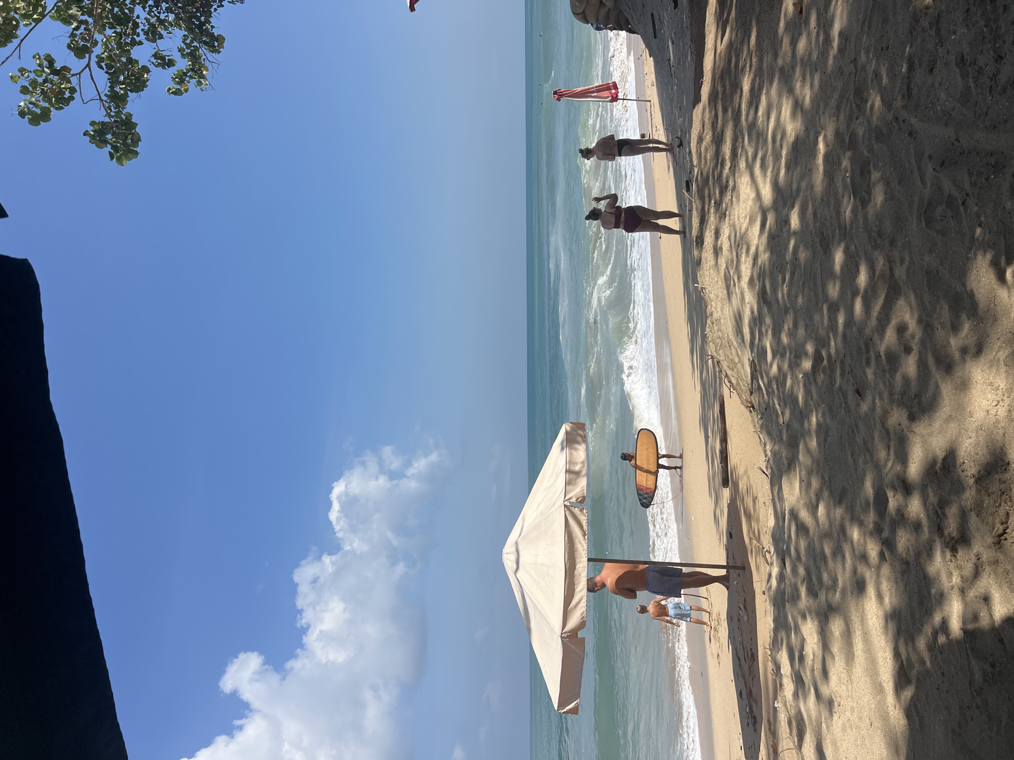 In Kuta, Indonesia, a sunny day at the beach unfolds with gentle waves lapping the shore. A large, beige umbrella provides shade on the sand, where a man and a young boy stand near the water's edge. To the right, a person carries a surfboard, facing the ocean. Further down the beach, two women in swimsuits converse, with a red and white umbrella and sandbags nearby. The sea is a tranquil blue, with small waves rolling in under a clear sky dotted with fluffy white clouds. The foreground shows the sandy beach with dappled sunlight filtering through tree shadows.