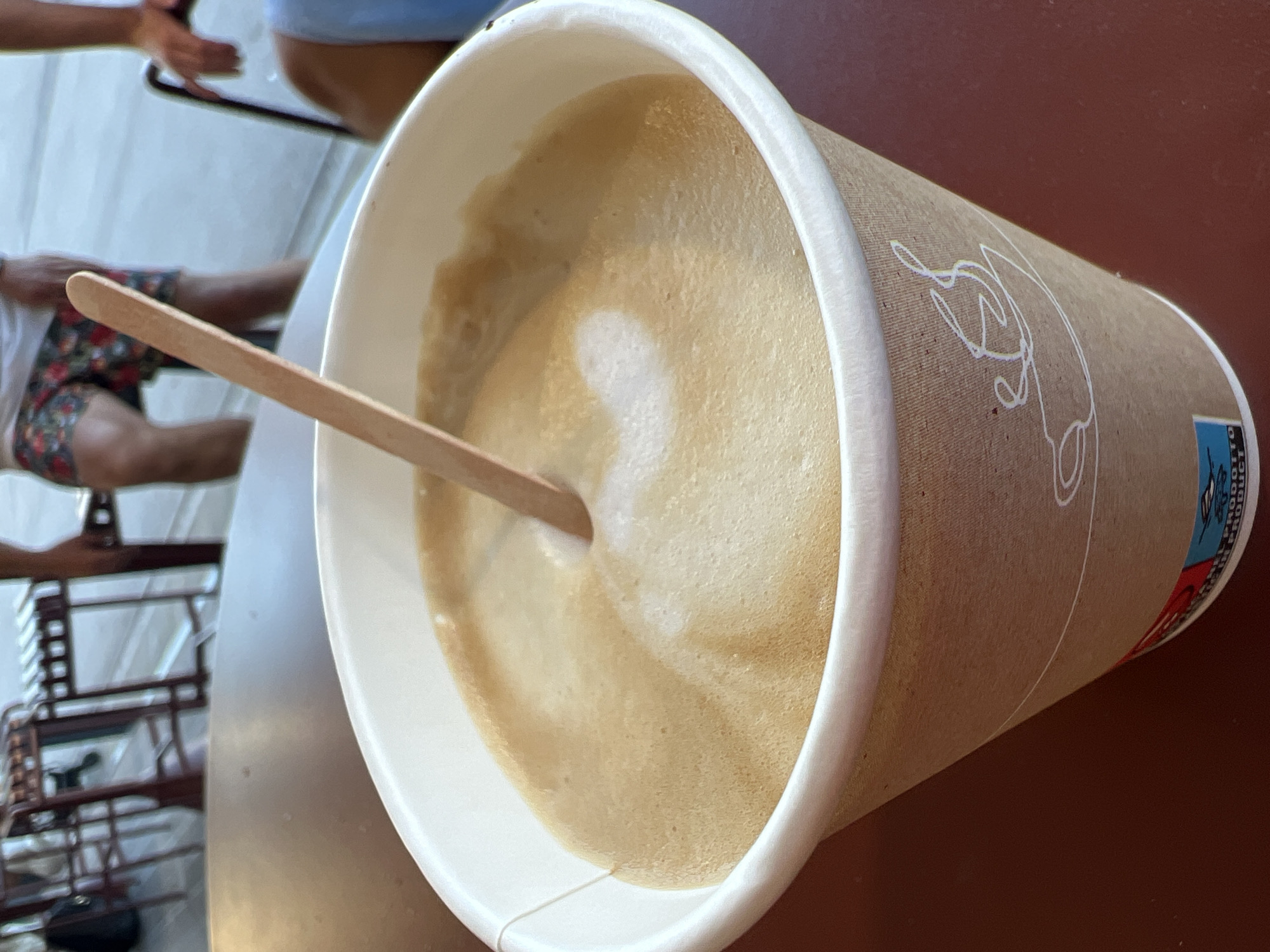 In Budva, Montenegro, a close-up shot focuses on a paper coffee cup filled with a frothy beverage, likely a cappuccino or latte. A wooden stir stick is partially submerged in the foam, creating a swirl pattern. The cup has a simple white rim and is adorned with a minimalist line drawing of a coffee cup. The background is blurred, showing hints of outdoor seating with red chairs and tables, suggesting a cafe setting. Partially visible in the upper right background are the legs of a person wearing floral-patterned shorts, and in the far right background, a person's hand rests on the back of a chair. The overall lighting suggests an outdoor daytime setting.