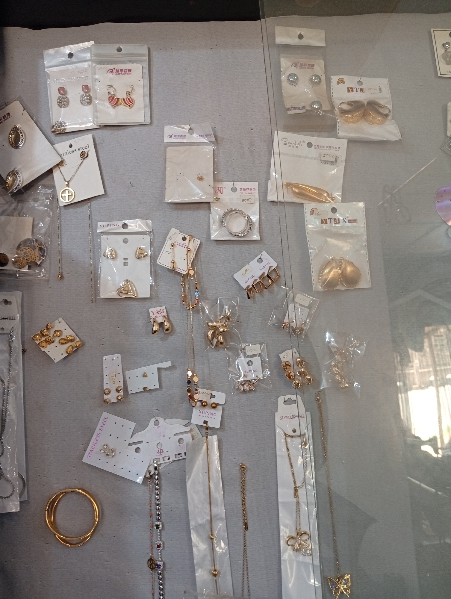 The image displays a close-up view of various pieces of jewelry, primarily earrings and necklaces, arranged on a gray, textured surface, likely a display shelf or wall. The items are mostly gold-toned and appear to be new, many still in their original packaging, which often features Chinese text and branding such as "XUPING" and "Y&M." Some individual pieces are also presented individually on cards.
Visible jewelry includes pairs of stud earrings with gemstones, dangling earrings with geometric and heart shapes, necklaces with pendants in the form of crosses, hearts, and bows, as well as bracelets and bangles. The lighting suggests an indoor setting, possibly a retail environment, with some reflections from a glass display case visible. The overall impression is of a well-organized display of fashion accessories. The question of its location in Jos, Nigeria, is not directly answerable from the visual information provided.