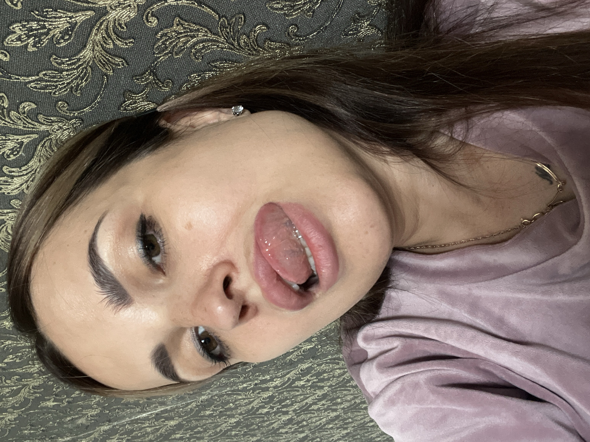 A young woman with brown hair and makeup is looking at the camera with her tongue sticking out. She is wearing a pink velour shirt and a gold chain necklace. She has a small diamond earring in her right ear. The background is a dark gray wallpaper with a gold floral pattern. The image is a close-up shot of the woman's face and upper body.