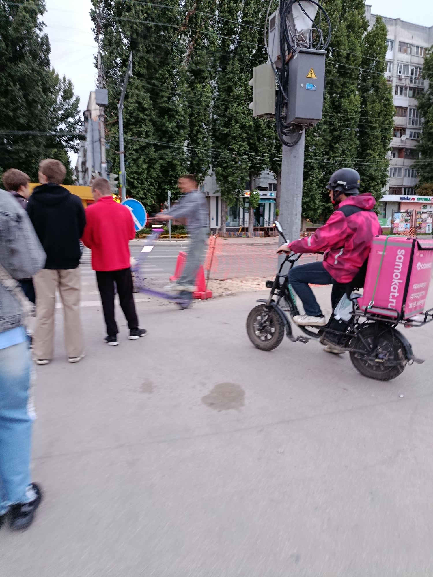 The image shows a street scene in Saratov, Russia, with several people and a person on an electric scooter. In the foreground, a person wearing a bright pink jacket with a "samokat.ru" logo on a thermal bag is riding an electric scooter. They are wearing a helmet and blue jeans. To the left, three people are standing on the sidewalk, facing away from the camera. One person is wearing a black hoodie, another a red sweatshirt, and a third person is partially visible in the foreground wearing a denim jacket. In the background, a utility pole with electrical equipment is visible, along with tall trees and apartment buildings. The street appears to be under construction or undergoing maintenance, with orange cones and barriers visible. The overall impression is a candid snapshot of urban life in Saratov.
