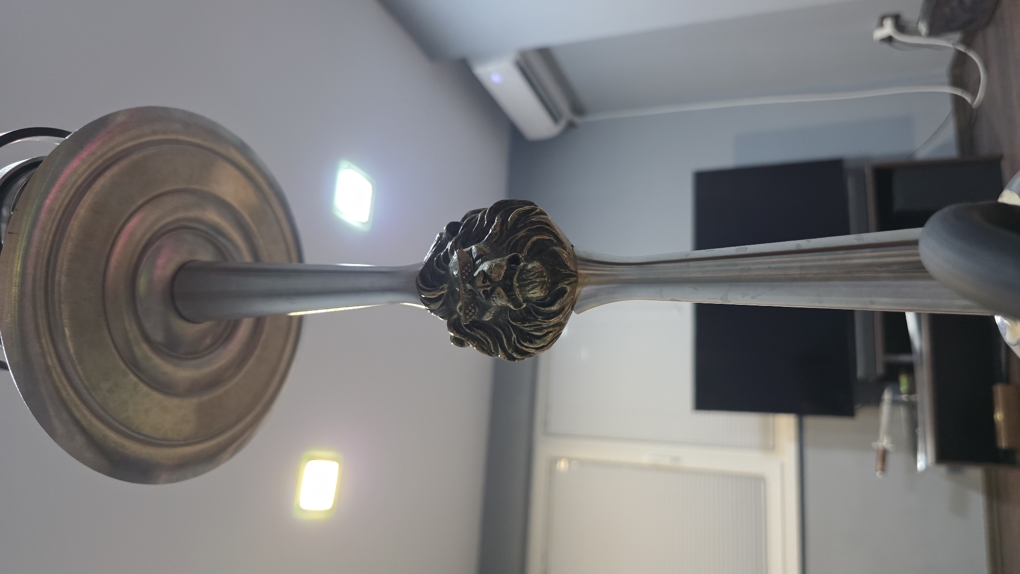 A low-angle, close-up shot focuses on the ornate head of a hookah stem, featuring a detailed lion's head motif with sunglasses. The stem, made of polished metal, extends upwards and is capped by a decorative, concentric-ringed disc. The background is an indoor environment, likely a room, with a plain gray wall, a white air conditioning unit mounted high up, and a dark television screen visible in the mid-ground. Two square, illuminated ceiling lights are also present. The floor is not clearly visible. The scene is devoid of human presence and appears to be a still life composition. The location is in Montana, Bulgaria.