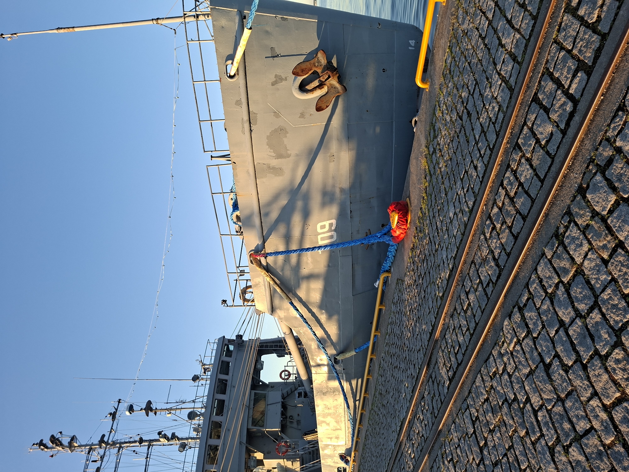 A grey naval or auxiliary ship, identified as EML Grif (A432) and displaying the hull number "09" on its starboard bow, is securely docked at Lennusadam (Seaplane Harbour) in Tallinn, Estonia. The ship's bow is visible in the foreground, featuring a large, distinctive anchor. Multiple mooring lines, varying in color (blue, yellow, and multi-colored), connect the vessel to the pier. A prominent blue mooring line is wrapped around a yellow bollard or cleat on the pier, with a red/orange fender or buoy positioned on the cobblestones nearby.
The pier surface consists of grey cobblestones, with two parallel, rusted metal rails embedded within them, indicating former trackage. Along the edge of the pier, a yellow metal barrier or railing is present. The ship's superstructure, including its bridge with visible windows and a mast equipped with various antennas and radar domes, is partially visible in the background. A cylindrical object, possibly a survey equipment davit or a small cannon, is discernible on the ship's port side, between mooring lines.
The sky above is clear and uniformly blue. The scene is illuminated by the low-angle, golden light of either sunrise or sunset, casting long shadows from the ship onto its own hull and across the cobblestone pier. The water in the harbor appears calm and blue. No individuals are visible in the image.