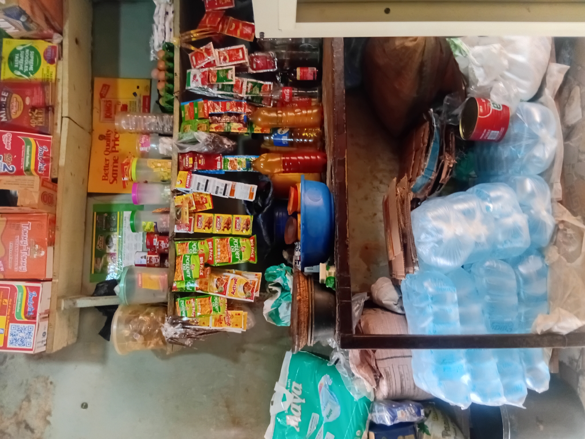 This image depicts a shelf inside a small shop or stall in Nkpor, Nigeria. The shelves are stocked with various items, including packaged foods, bottles of drinks, and bags of groceries. On the top shelf, there are boxes of noodles, a tin labeled "MIKEE," and bags of green food items. Below that, on the middle shelf, are several plastic containers, a tray of eggs, and numerous small sachets of spices and condiments hanging on display. To the left, a large blue plastic bag labeled "Maya" is visible, possibly containing diapers or sanitary products. The bottom shelf holds stacks of bottled water, a red tin can, and other assorted items covered in plastic. The overall environment appears to be a retail setting, likely a local convenience store or market stall, with a cluttered but organized arrangement of goods. The lighting suggests an indoor environment, possibly with natural light filtering in from an unseen source.