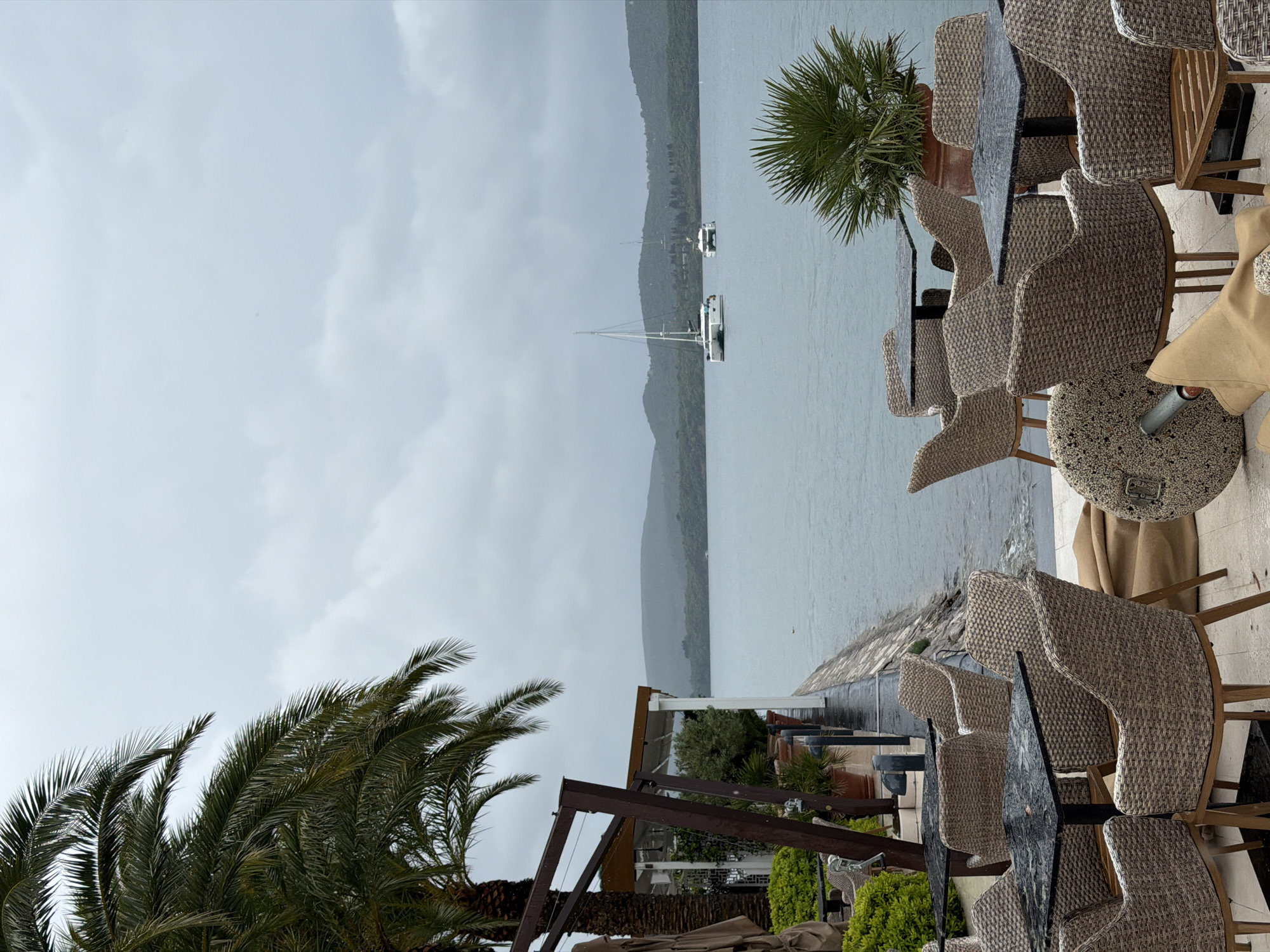 The image shows an outdoor seating area, likely a restaurant or cafe, in Tivat, Montenegro. Wicker chairs and tables are arranged overlooking a body of water. In the background are hills or mountains, and several boats are visible on the water. The sky is overcast and gray, suggesting a rainy or cloudy day. Palm trees are prominent in the foreground, adding a tropical feel to the scene. The overall atmosphere is calm and tranquil, despite the weather. No people are visible in the image.