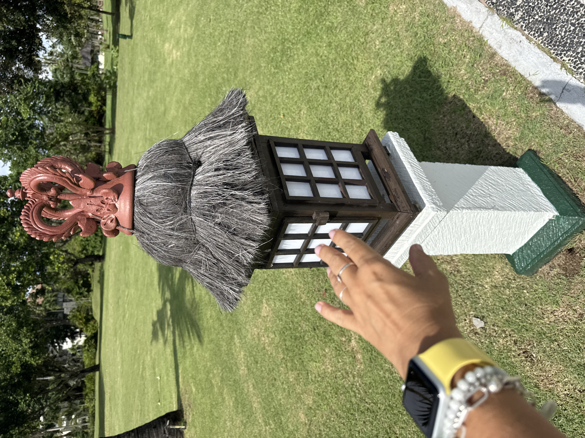 The image, oriented incorrectly and showing a horizontal view of a vertical scene, depicts a person's hand reaching towards a decorative outdoor lantern positioned on a well-maintained grassy area.
In the foreground, a light-skinned hand with a silver ring on the ring finger extends towards the lantern. The person's wrist wears a bright yellow watch band and a beaded bracelet with multiple silver-colored charms.
The prominent object is an ornate lantern or lamp post. It features a dark wooden body with multiple square, opaque white panels, suggesting it is a light fixture. Above the main lamp section, there's a layered, textured covering resembling thatch or dried grass, giving it a rustic or traditional appearance. Crowning the entire structure is a reddish-brown, intricately carved finial, possibly depicting a mythical creature like a dragon or phoenix, with elaborate swirling patterns. The base of the lantern is a white concrete or stone pillar, with a distinct green border or trim near the ground.
The setting is an outdoor area, likely a garden, lawn, or park. A vast expanse of short, vibrant green grass stretches into the background. Several trees with dense green foliage are visible further back, indicating a lush environment. The bright sunlight casts distinct shadows on the grass and from the lantern, suggesting it is daytime with clear skies. On the far right, a paved path with small dark pebbles and a concrete border separates the grass from an unseen area.
No visible text is present in the image. The style of the lantern, particularly the carving and thatched element, aligns with the provided location context of Kuta Selatan, Indonesia.