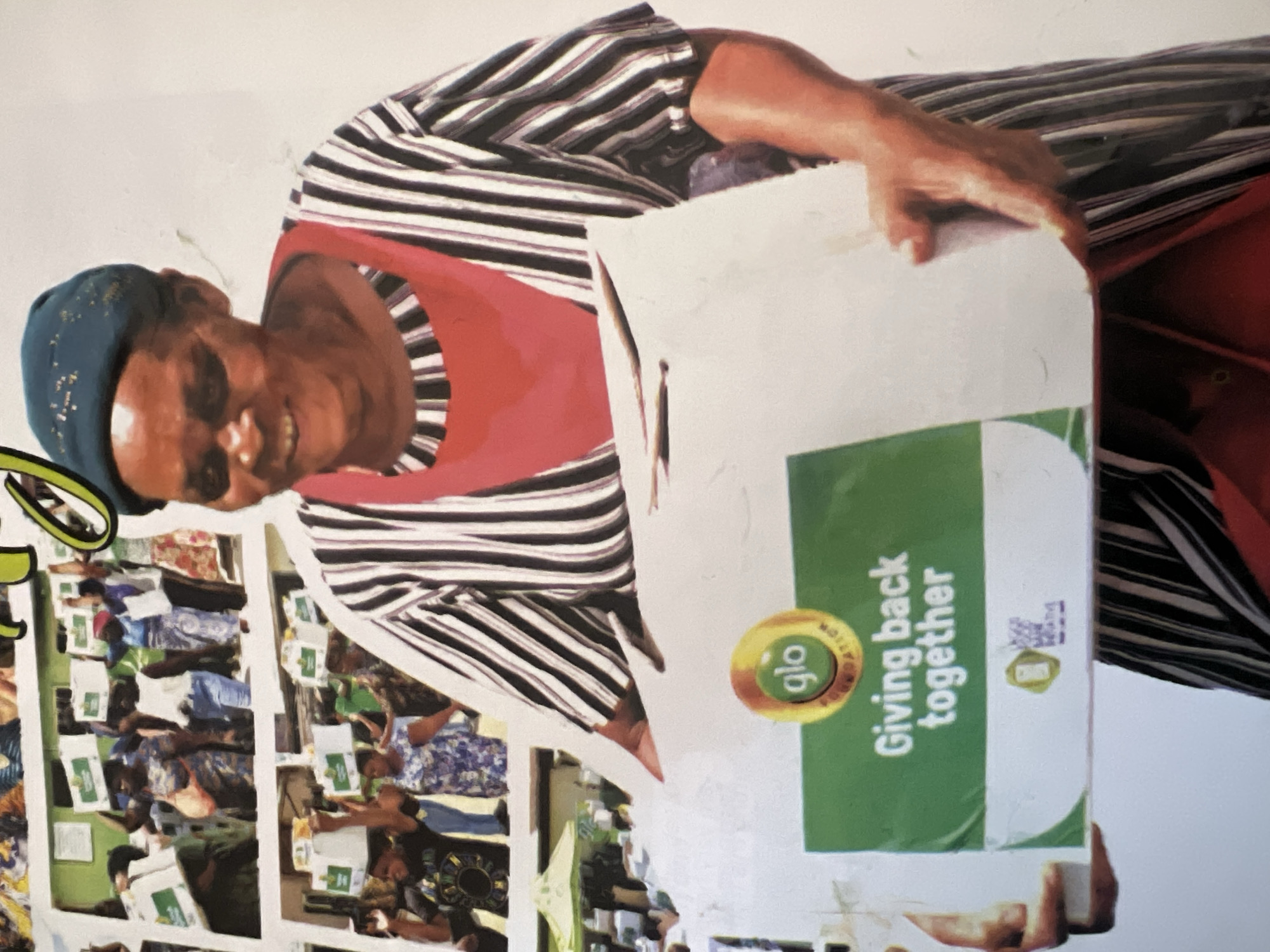 The image shows a woman in Jalingo, Nigeria, holding a large white box with green and white text. The text on the box reads "glo FOUNDATION" and "Giving back together". The woman is wearing a dark blue headwrap and a black and white striped dress over a red apron. She is smiling and looking to her right. In the background, there are several other people also holding boxes. Some are walking in a line, and others are standing. There are also some tents visible in the background, suggesting an outdoor event. The scene appears to be a community event or distribution of supplies.
